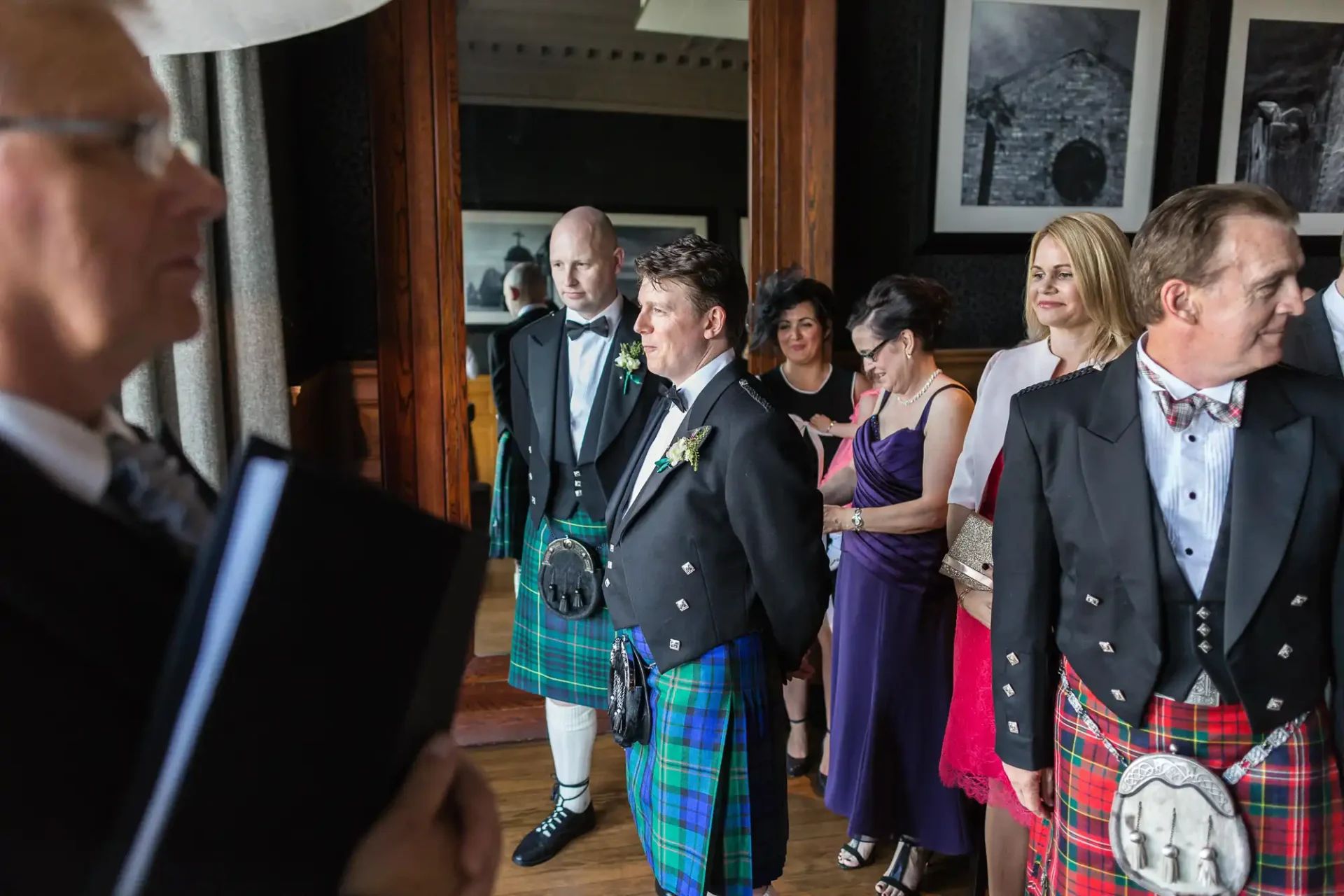 A group of people in formal attire, including kilts, attentively listening to a speaker in a room with traditional decor.