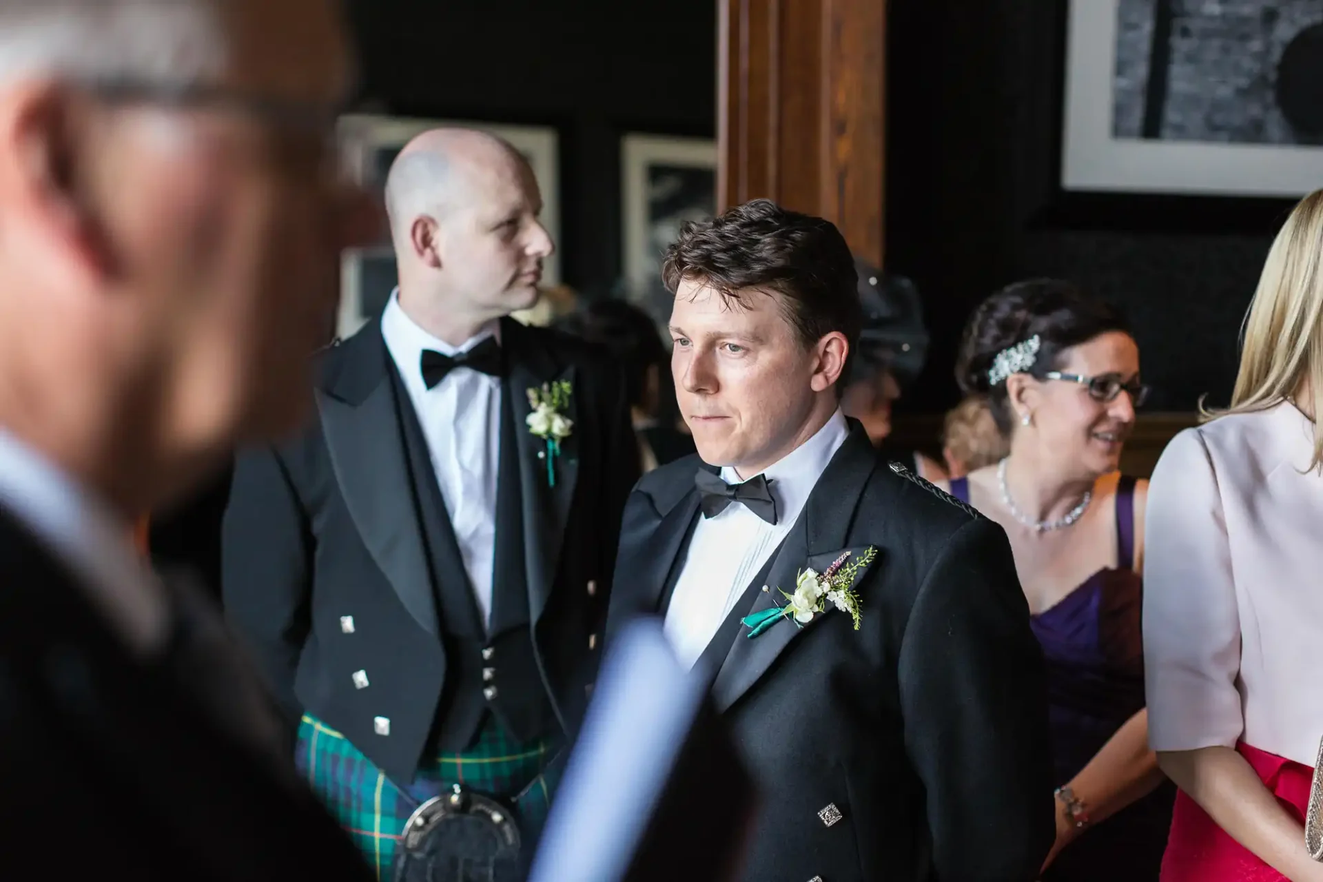 Groom in a black tuxedo looks thoughtful at a wedding reception, surrounded by guests including a man in a kilt.