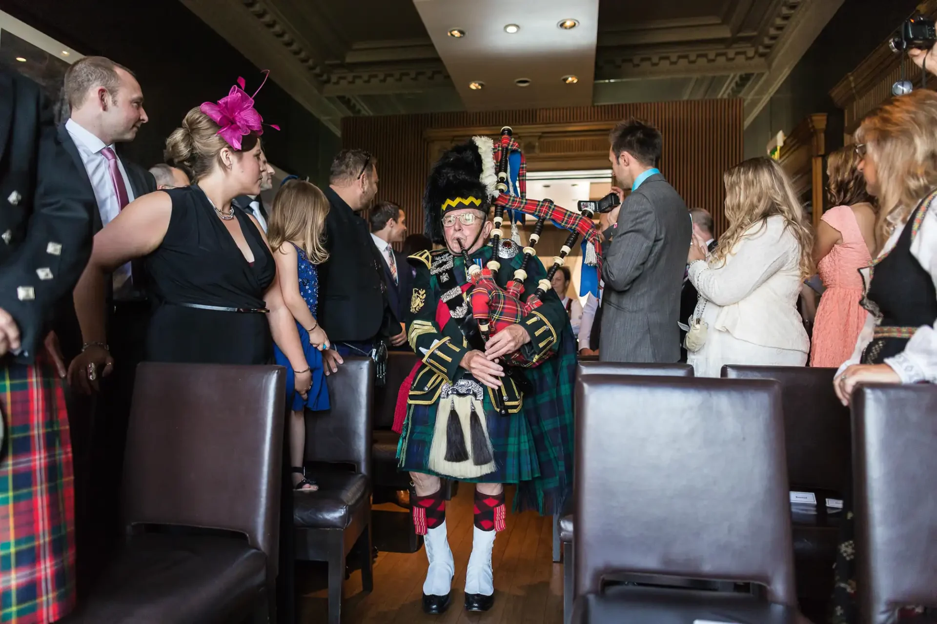 A bagpiper in traditional scottish attire plays inside a bustling room while guests in formal wear listen.