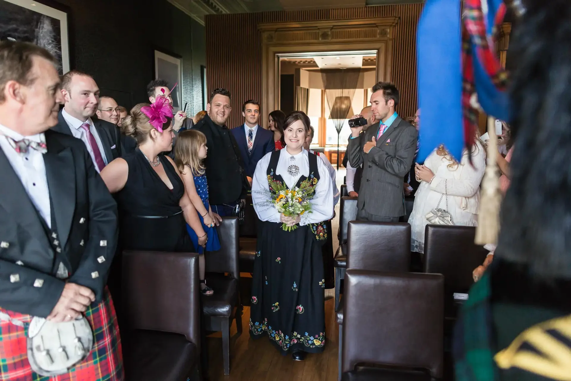 Woman in traditional dress holding a bouquet walks down the aisle at a wedding ceremony, surrounded by guests in various outfits, including kilts.