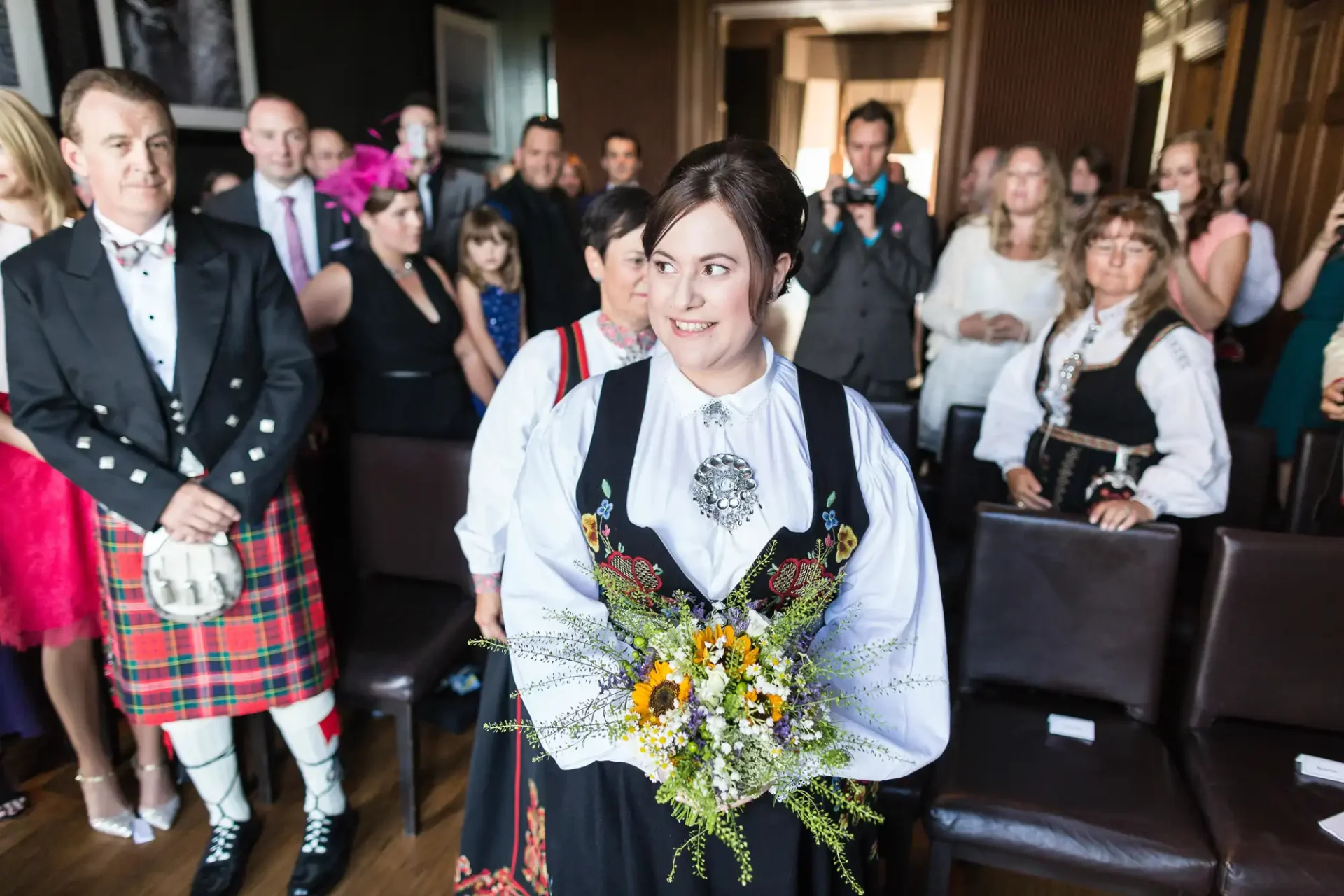 A bride in a traditional norwegian dress holding a bouquet, surrounded by guests in diverse attire at a wedding ceremony.