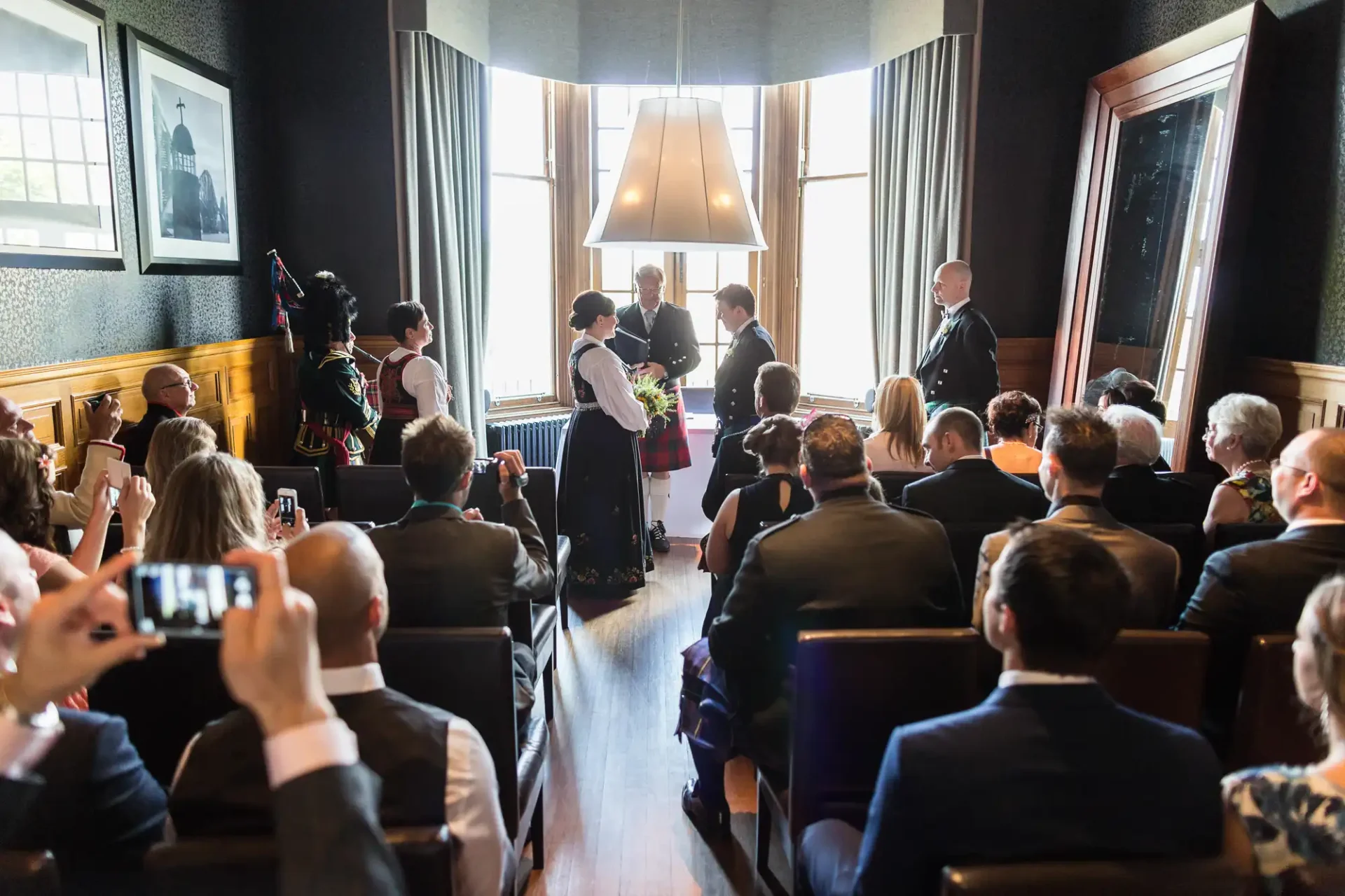 Wedding ceremony in a room with guests watching; bride and groom at the altar with an officiant and a bagpiper in attendance.
