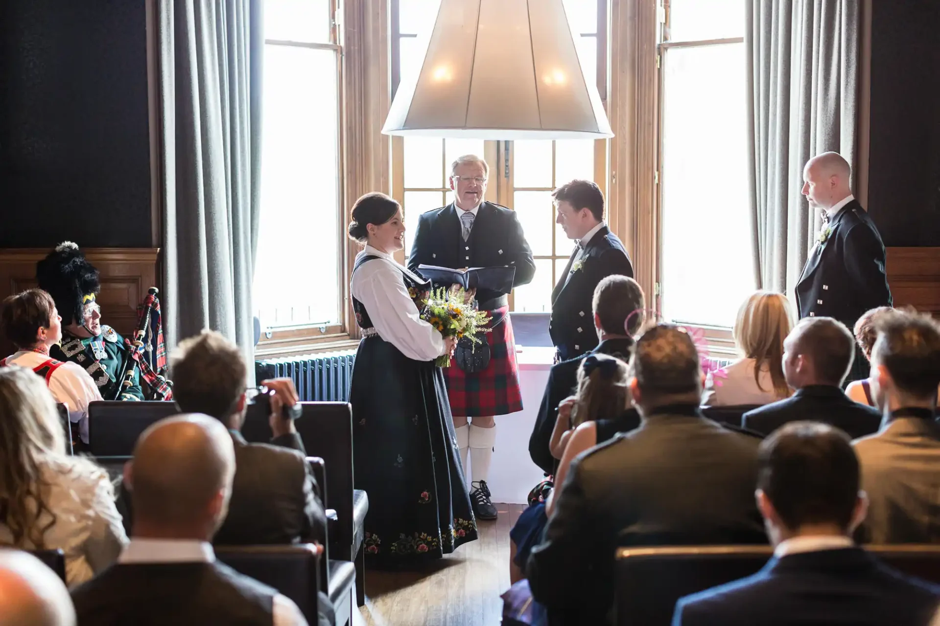 A wedding ceremony in a well-lit room featuring a bride and groom, the groom in a tartan kilt, with a bagpiper and seated guests watching.