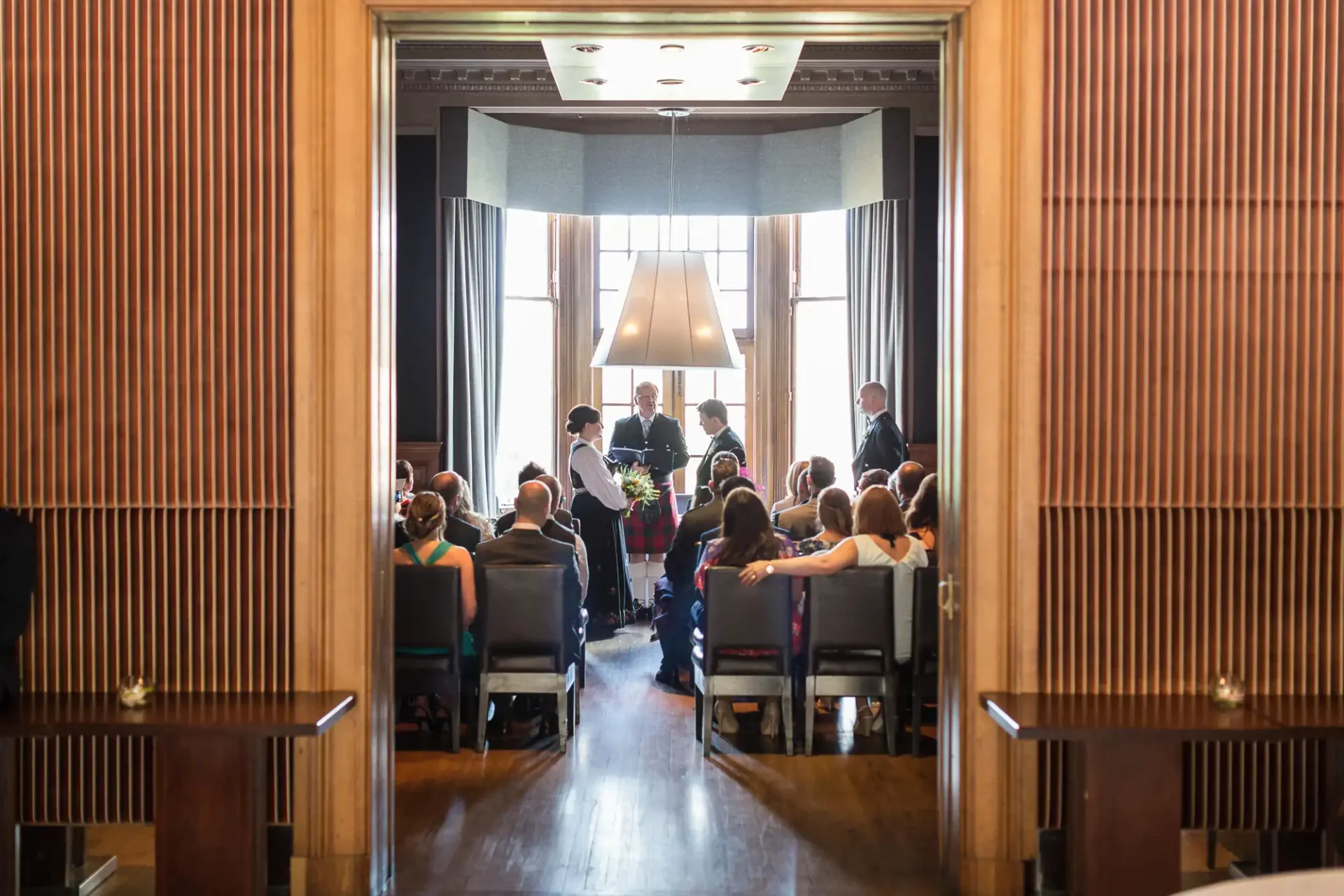 A wedding ceremony in a wood-paneled hall with guests seated as the couple stands before the officiant.
