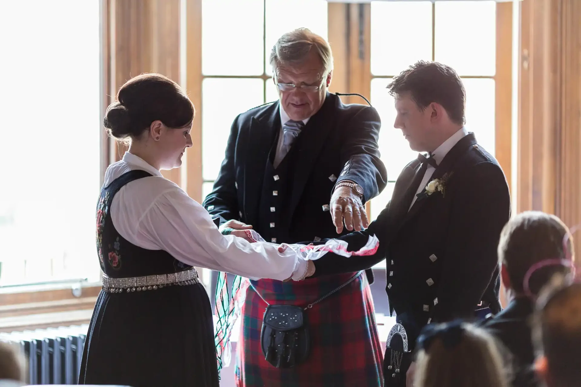 A wedding officiant conducts a handfasting ceremony for a bride and groom dressed in traditional scottish attire.