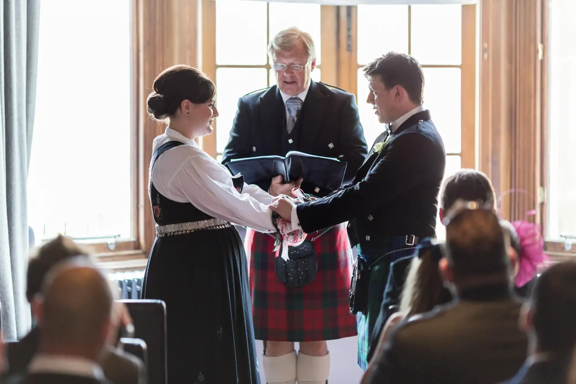 A wedding ceremony inside a room with a couple dressed in traditional scottish attire, exchanging vows while holding hands, with an officiant and guests in the background.