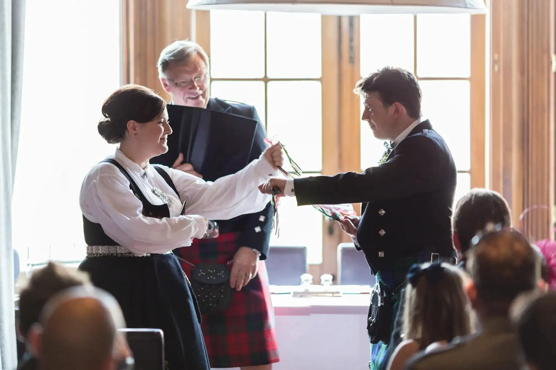 A bride in a traditional dress and a groom in a kilt exchanging rings at a wedding ceremony with an officiant and guests in the background.