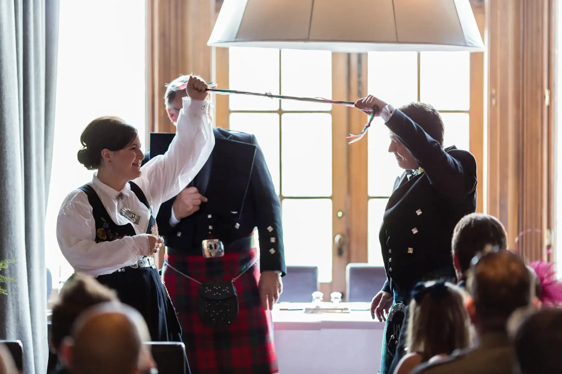 A man in a kilt and a woman in a black jacket holding a ceremonial sword over a newlywed couple at a scottish wedding.