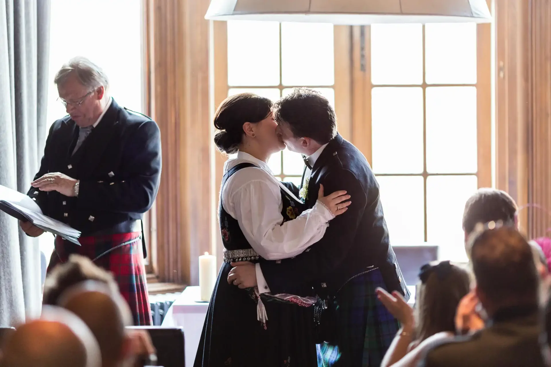 A couple in traditional scottish attire kissing at their wedding, with an officiant and guests in the background.