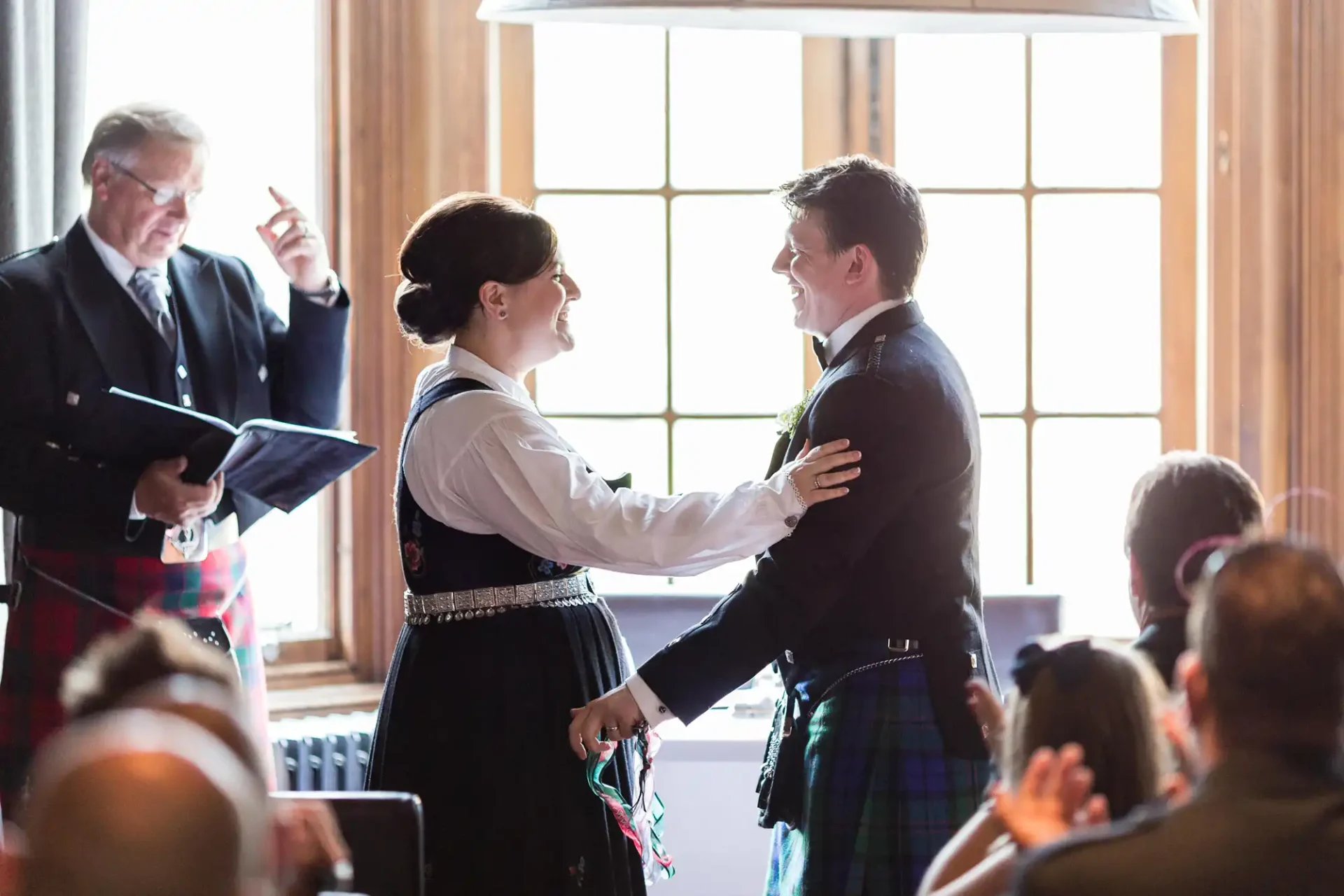 A bride and groom in traditional scottish attire exchange vows at a wedding ceremony, with an officiant and guests in the background.