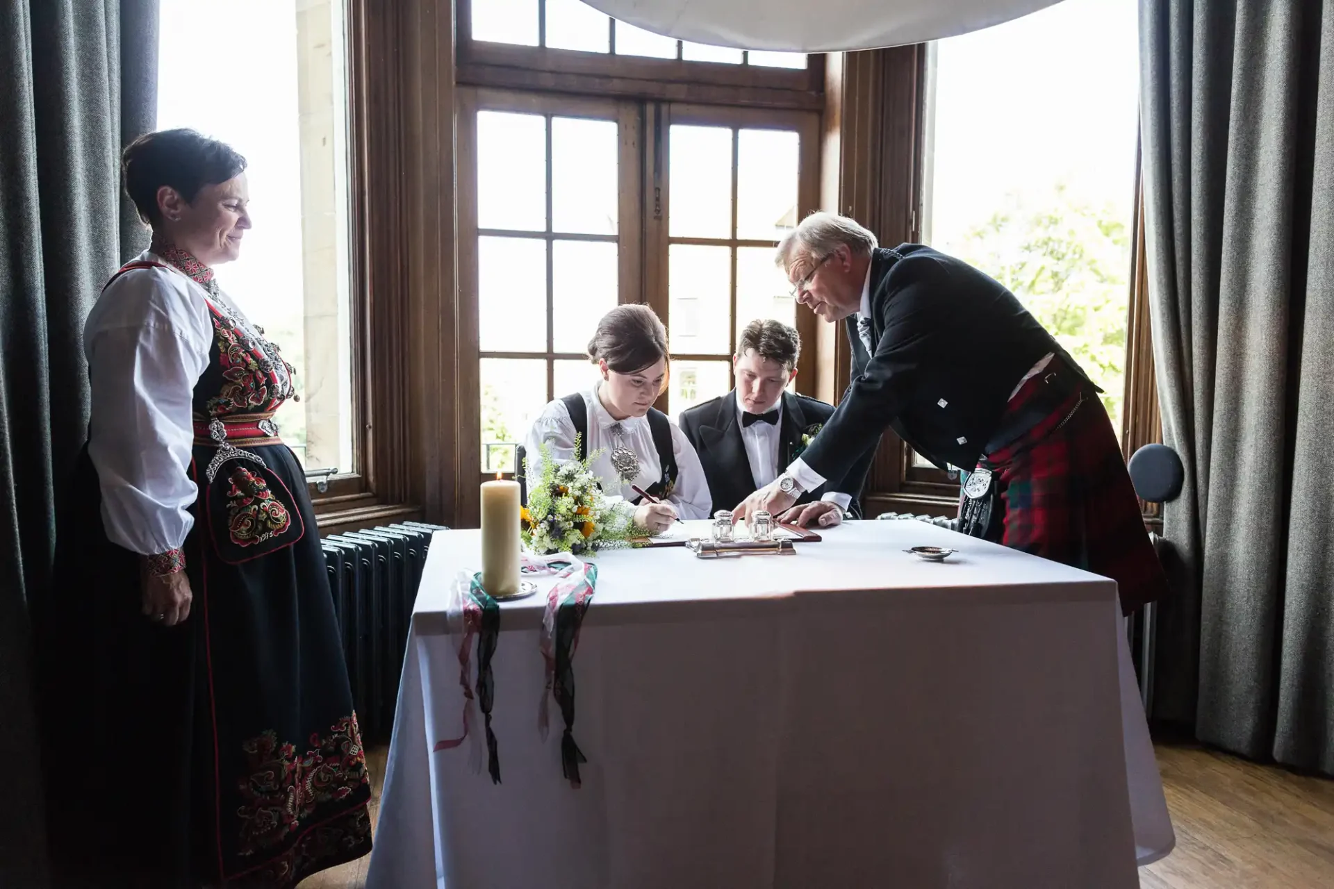 A wedding couple signs a register at a table, assisted by an official, inside a room with traditional décor and large windows, with two people in ethnic attire observing.