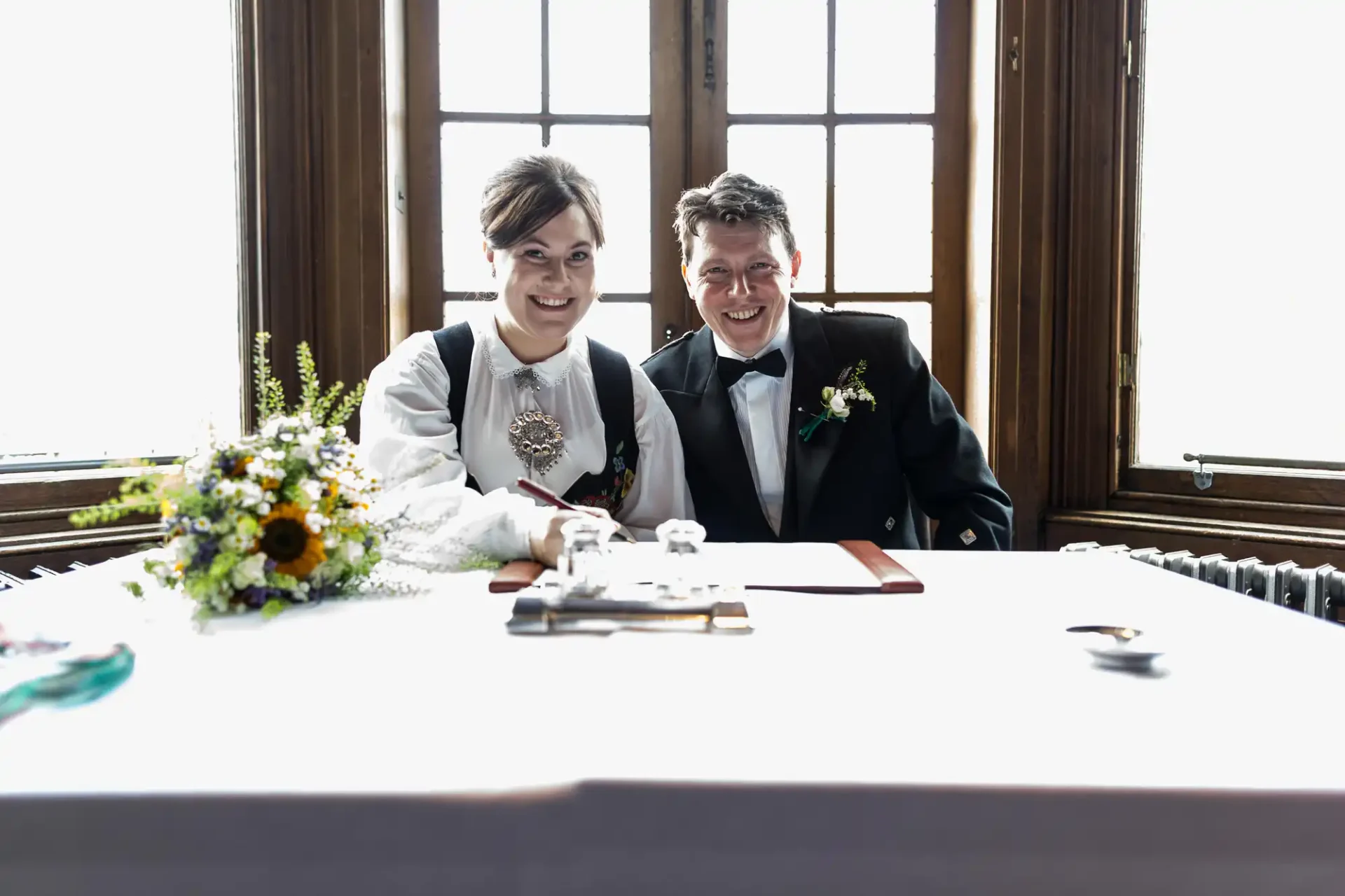 A bride and groom smiling at a signing table with a bouquet and documents, against a bright window background.