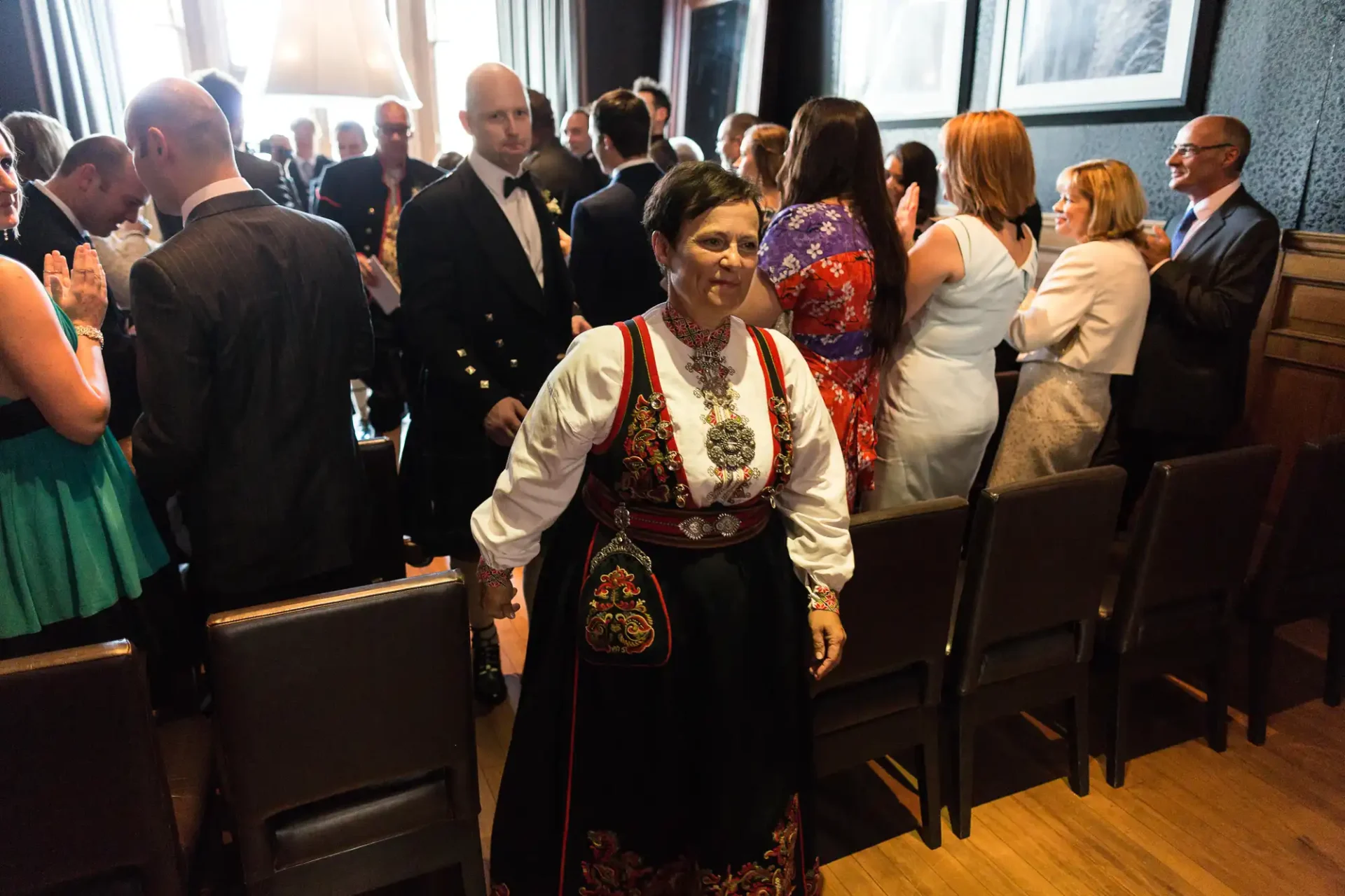 A woman in traditional norwegian dress at a crowded indoor event, smiling as she walks between rows of seated and standing guests.