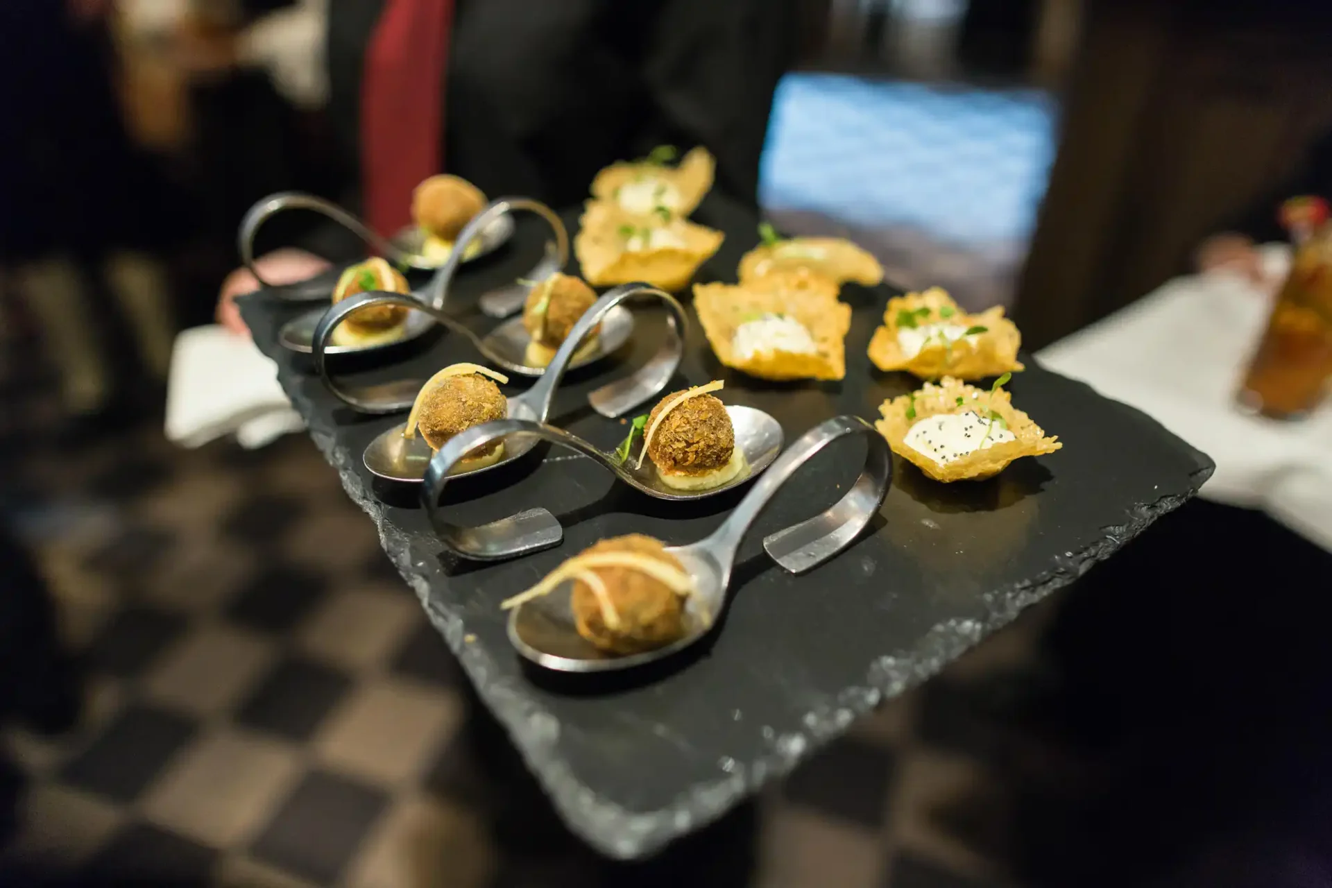 An array of assorted appetizers presented on modern spoons on a slate tray, held by a server in a restaurant.