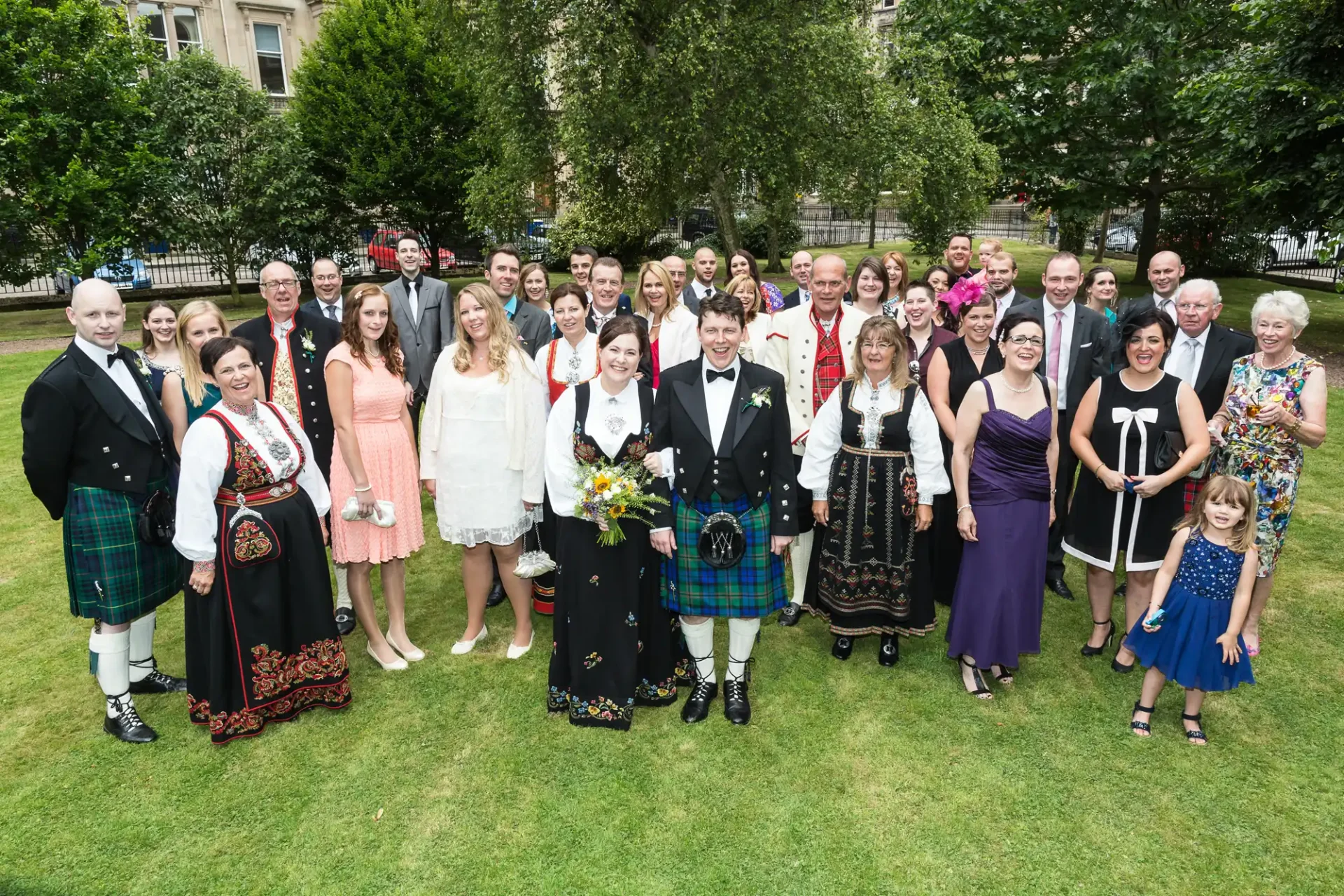 A group of people in formal attire, including kilts and suits, posing for a photo at a wedding outdoors.
