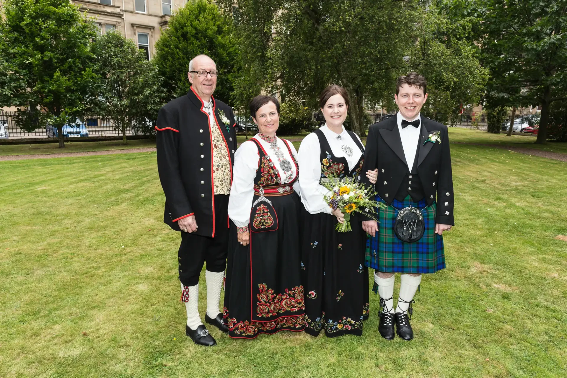 Two couples in traditional attire, one in a kilt and the other in norwegian dress, posing together in a green park.