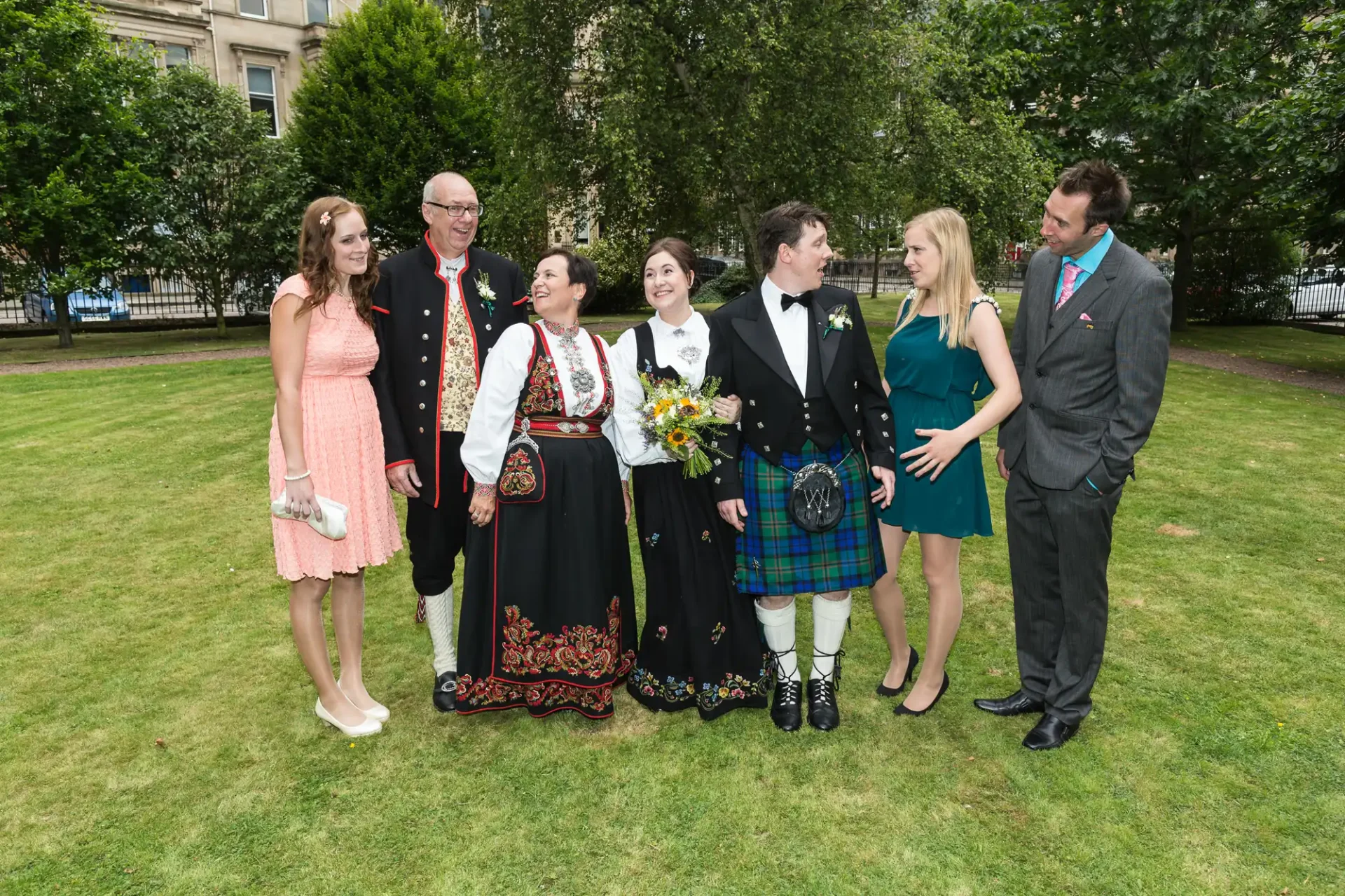 A group of seven people in formal attire, including kilts and a traditional norwegian dress, smiling together in a lush park.