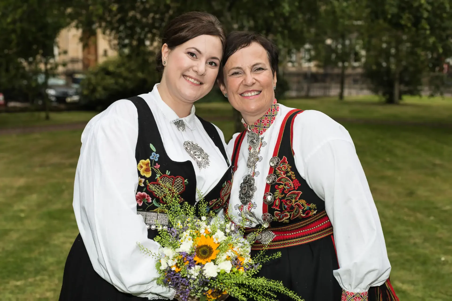 Two women in traditional norwegian dresses, holding a bouquet of wildflowers, smiling in a park.