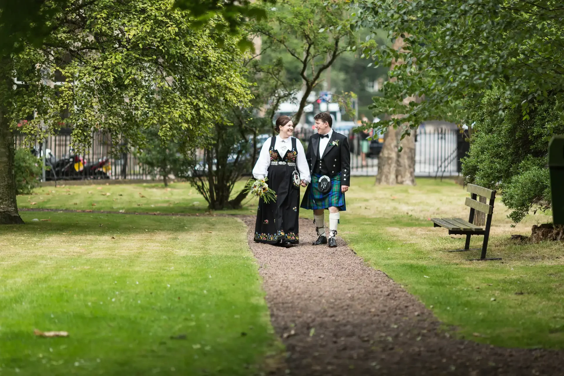 A couple in traditional scottish attire walking hand in hand along a park path, with trees and a bench in the background.