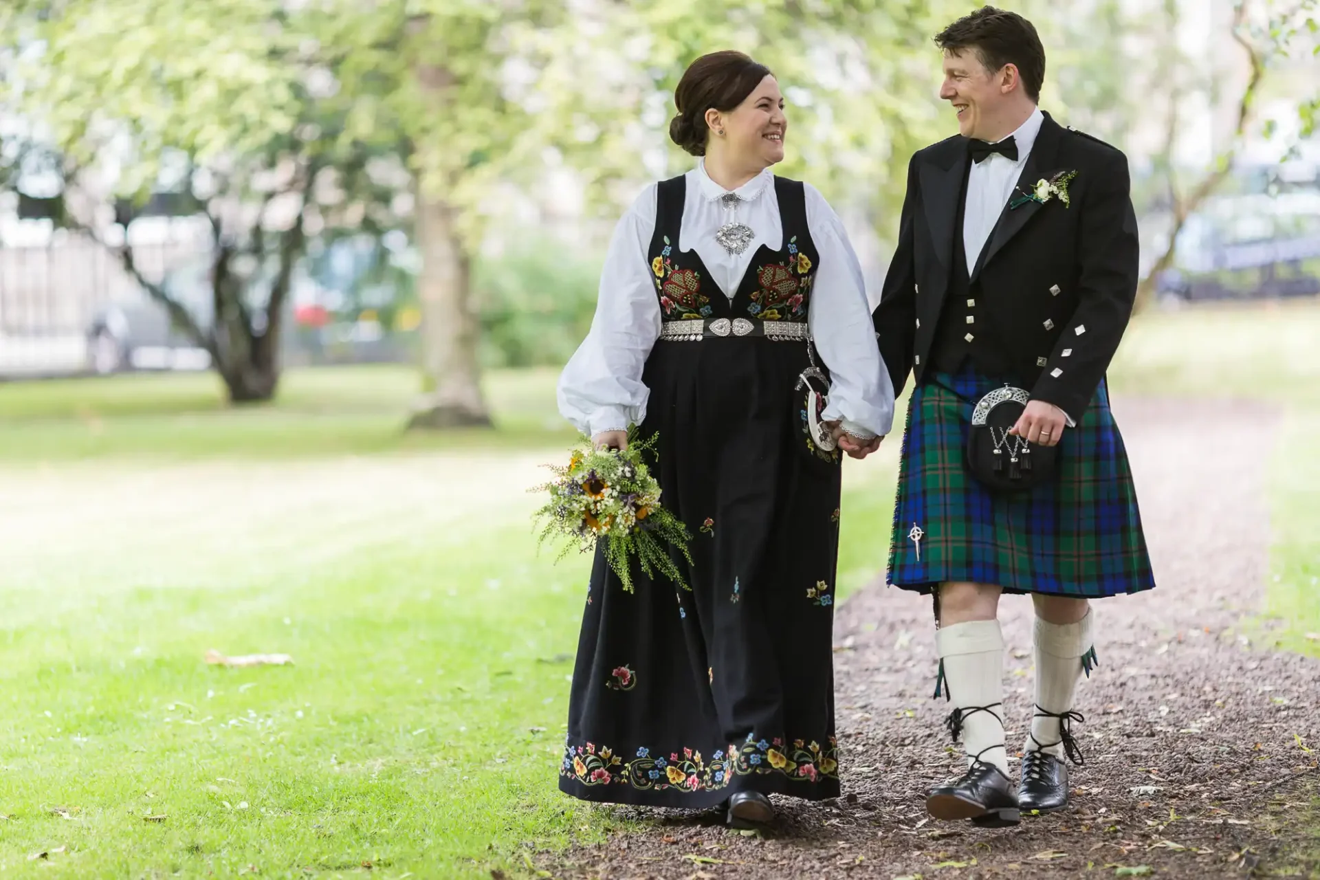 A bride in a traditional norwegian bunad and a groom in a scottish kilt holding hands and smiling at each other in a park.
