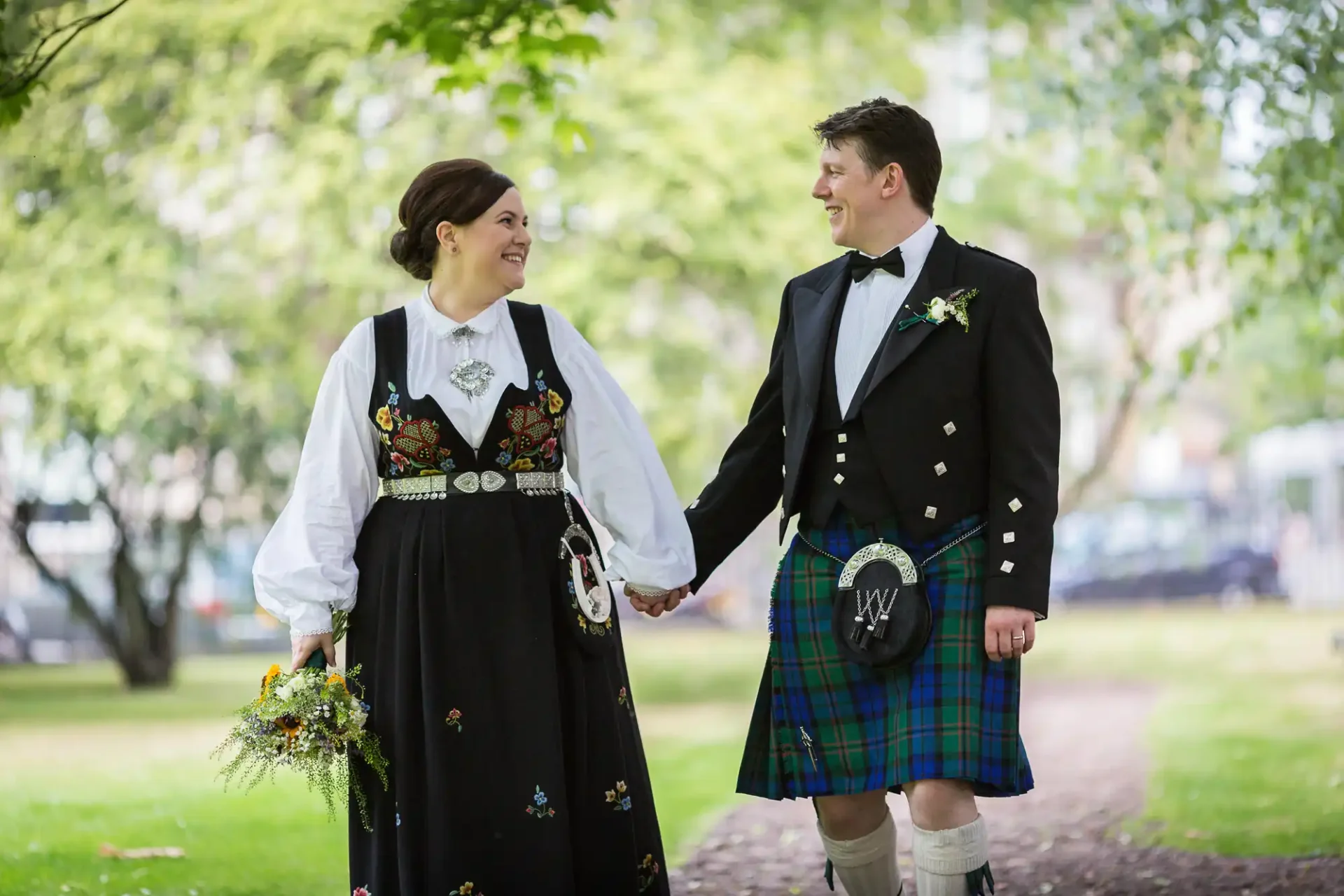 A bride in a traditional norwegian dress and a groom in a scottish kilt, holding hands and smiling at each other in a park.