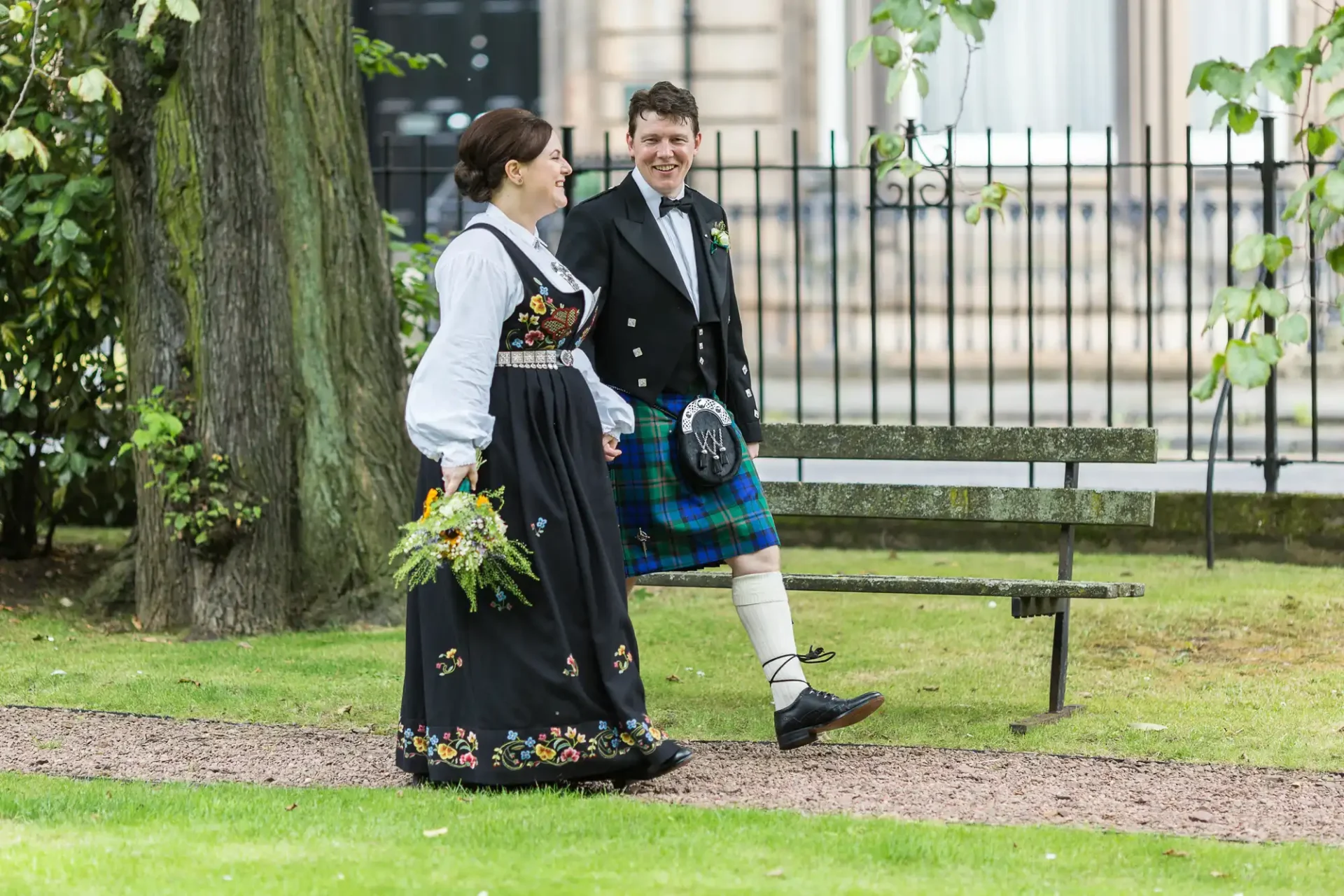 A couple in traditional attire walking in a park, the woman in a floral dress and the man in a tartan kilt, both smiling.