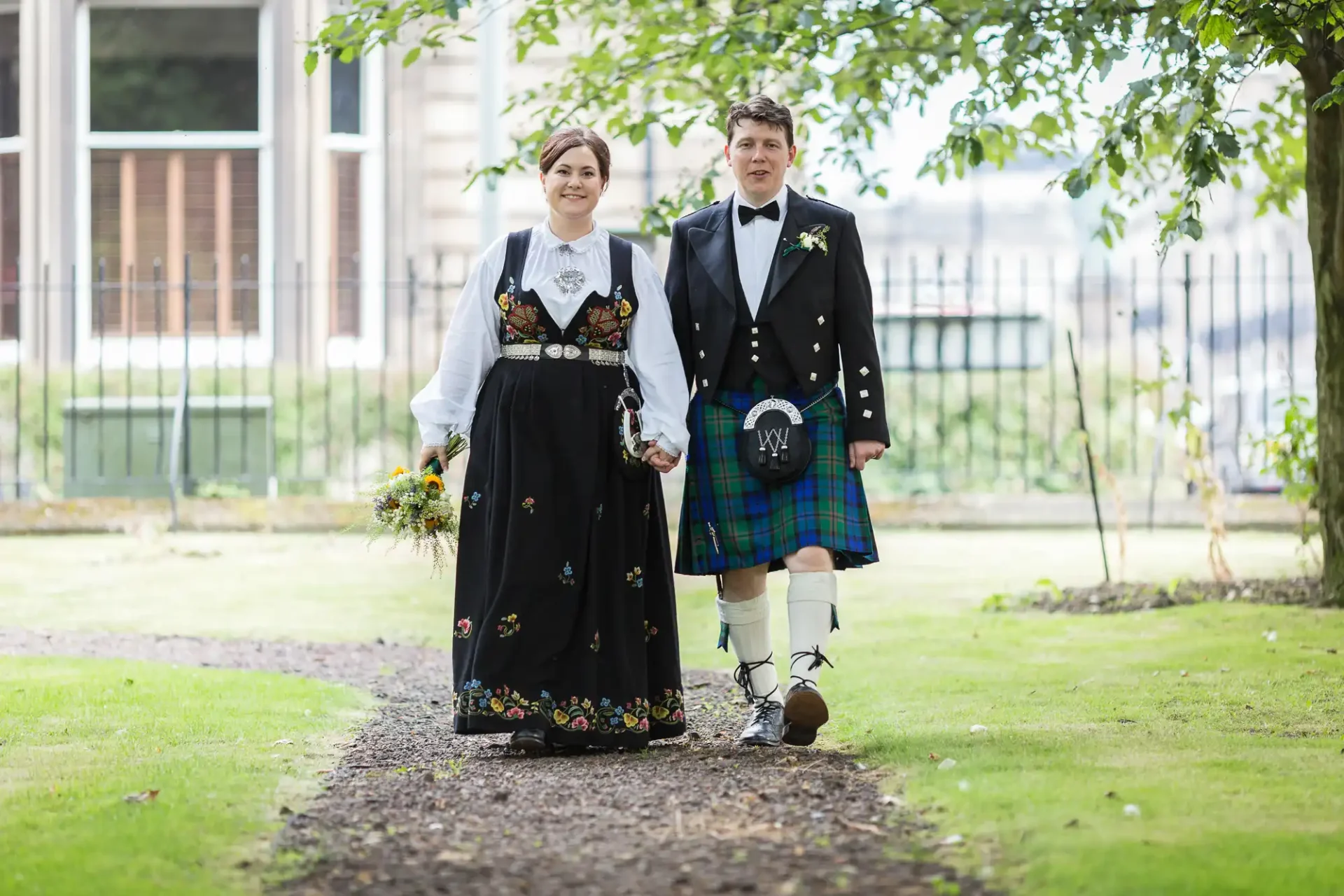 A couple in traditional norwegian and scottish attire walking hand in hand down a tree-lined path at their multicultural wedding.