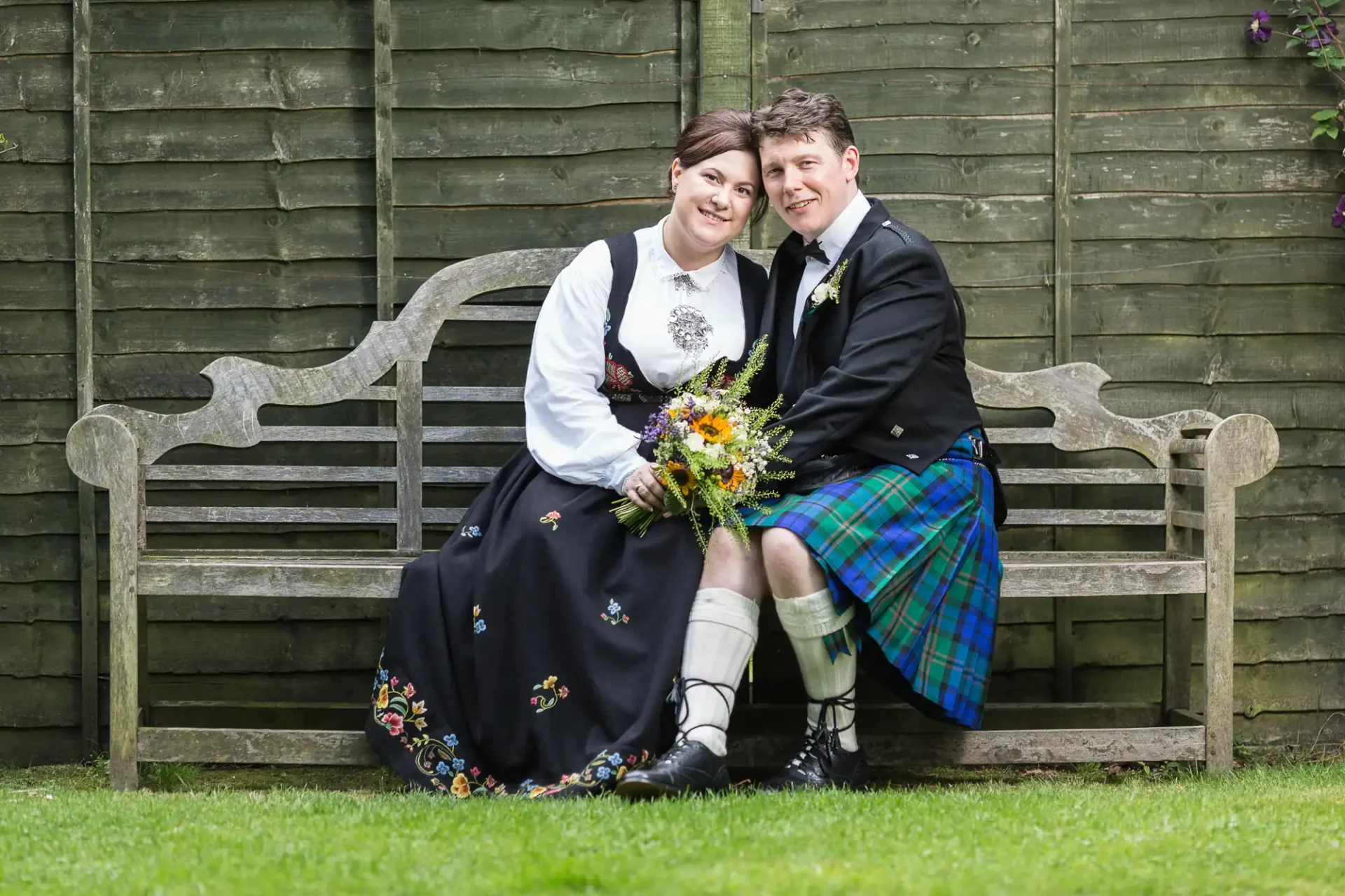 A couple in traditional scottish attire sitting closely on a garden bench, smiling at the camera, with a backdrop of a wooden fence and green grass.