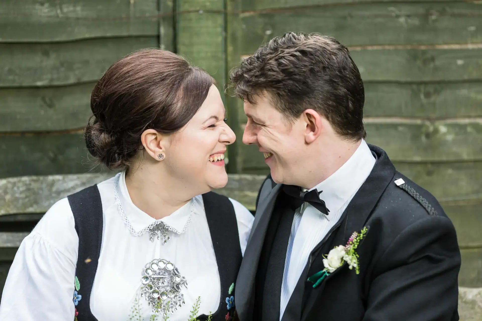 A joyful couple in traditional and formal attire smiling at each other with a wooden backdrop.