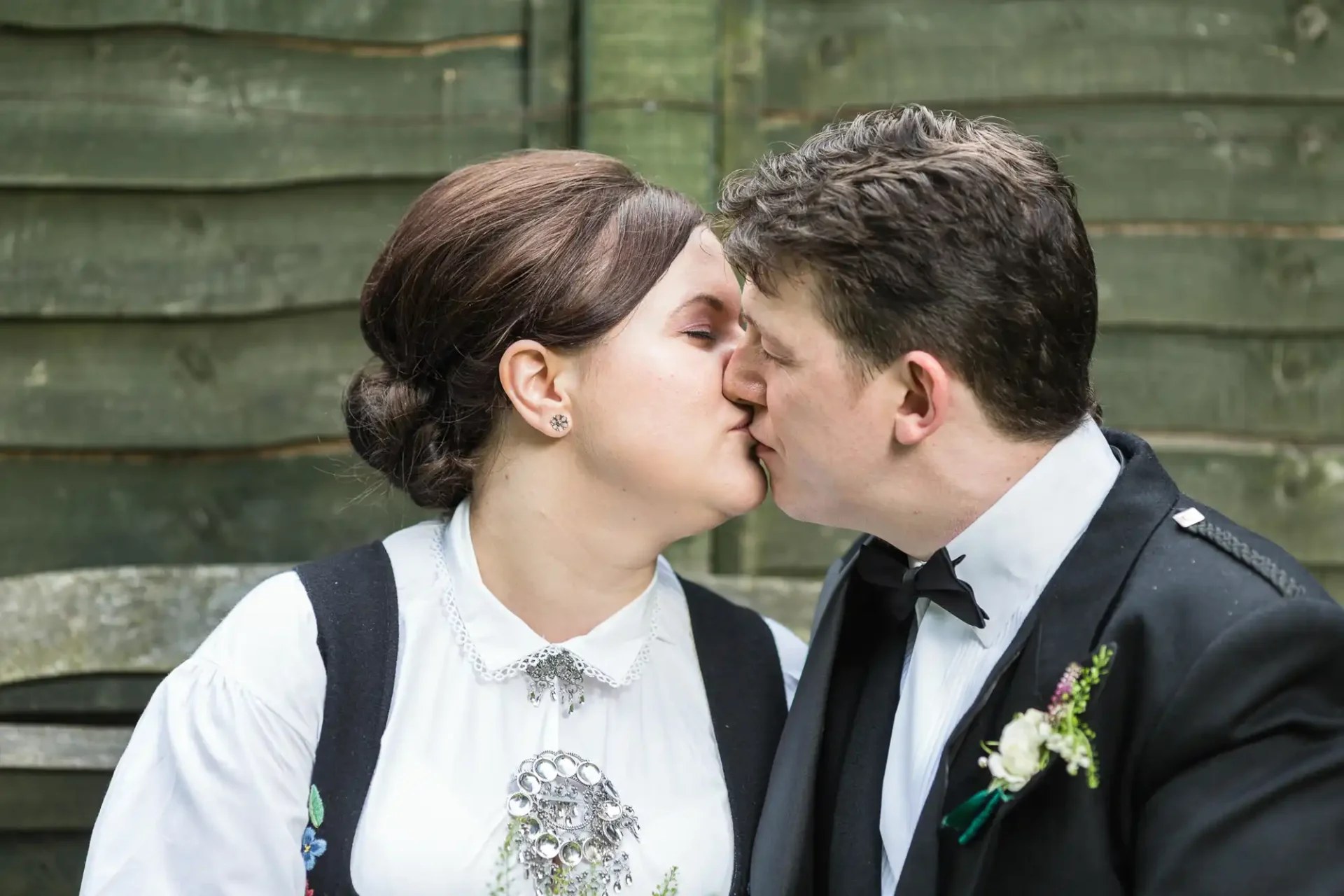 A couple in traditional attire kissing gently, with a wooden fence and greenery in the background.