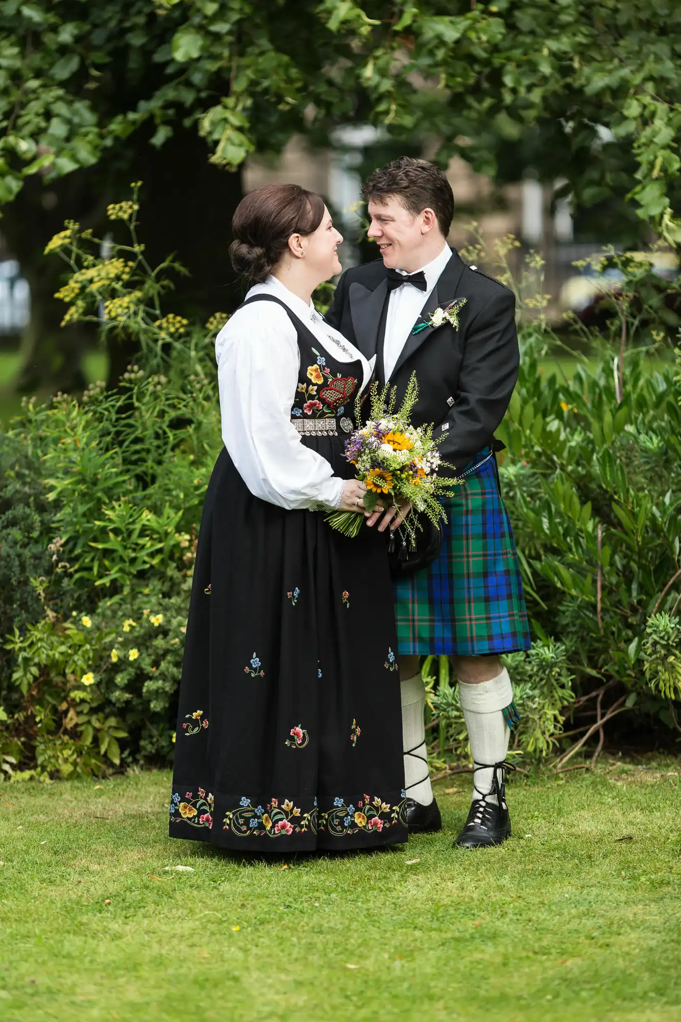 A couple in traditional scottish and norwegian attire smiling at each other in a lush garden, holding a bouquet.