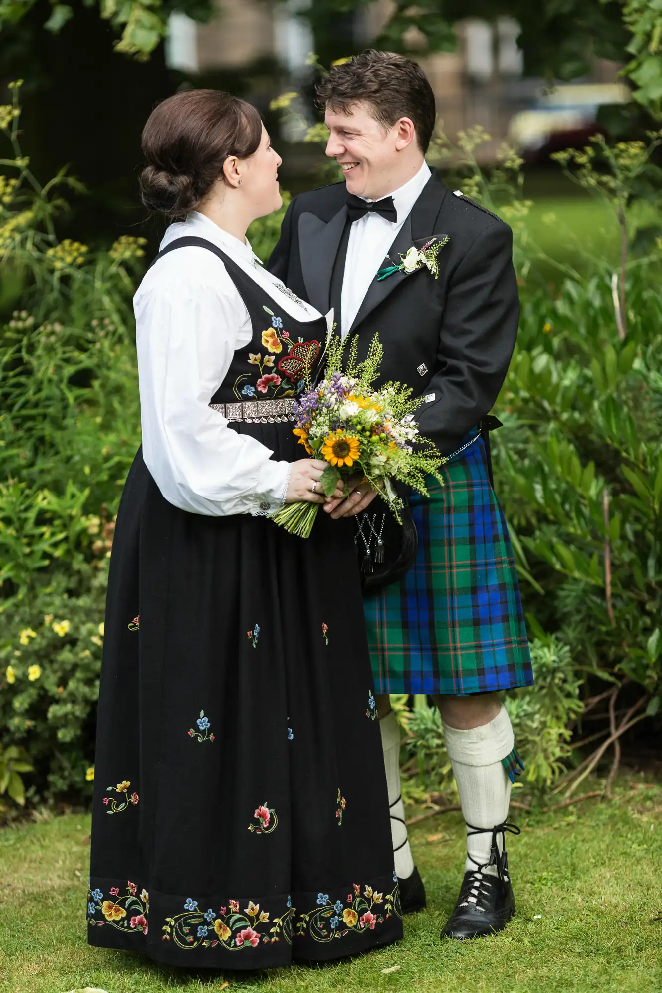 A bride in a traditional norwegian dress and a groom in a scottish kilt, smiling at each other and holding a bouquet, standing in a garden.