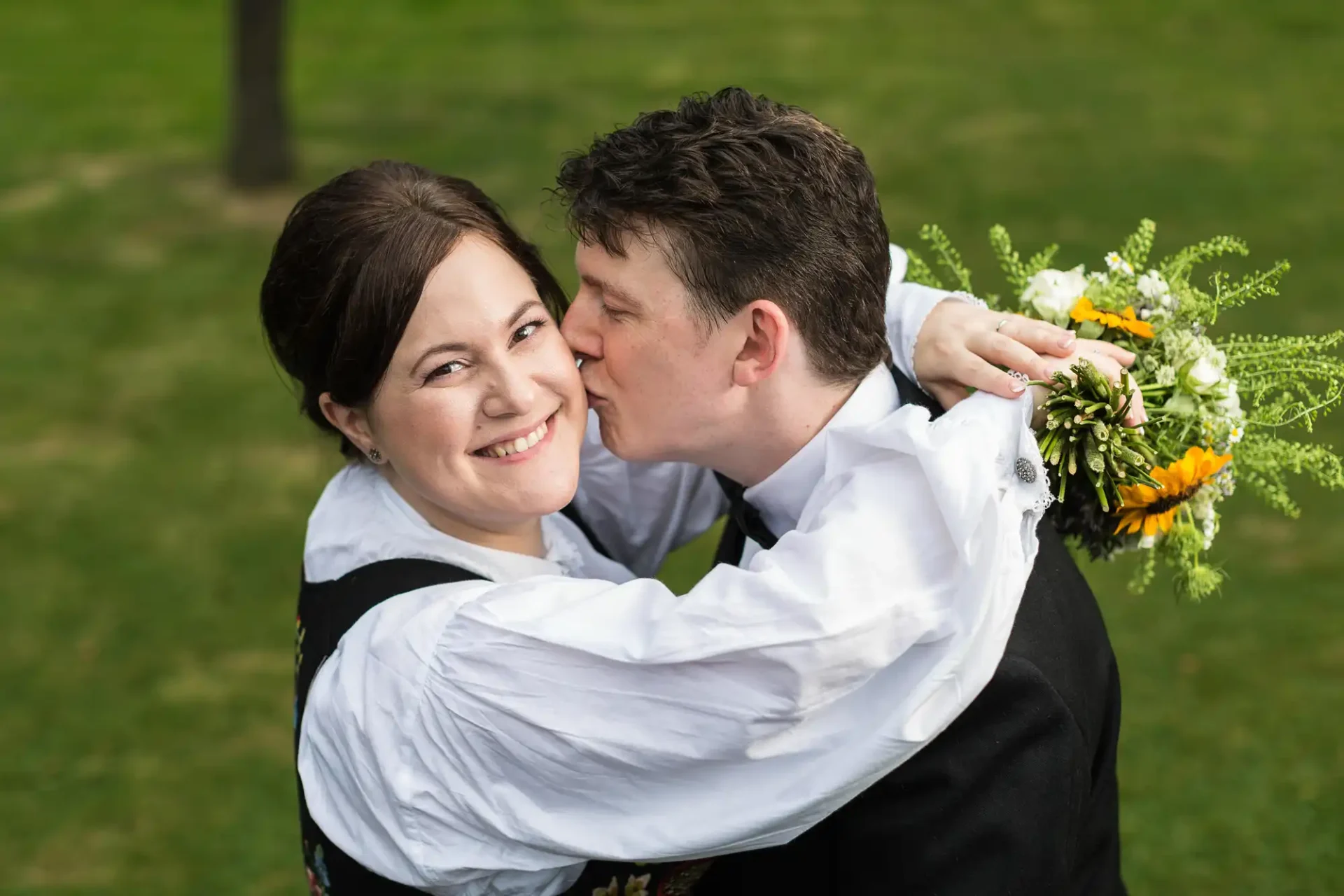 A couple in traditional attire embracing and smiling at a wedding, with the groom kissing the bride on the cheek.