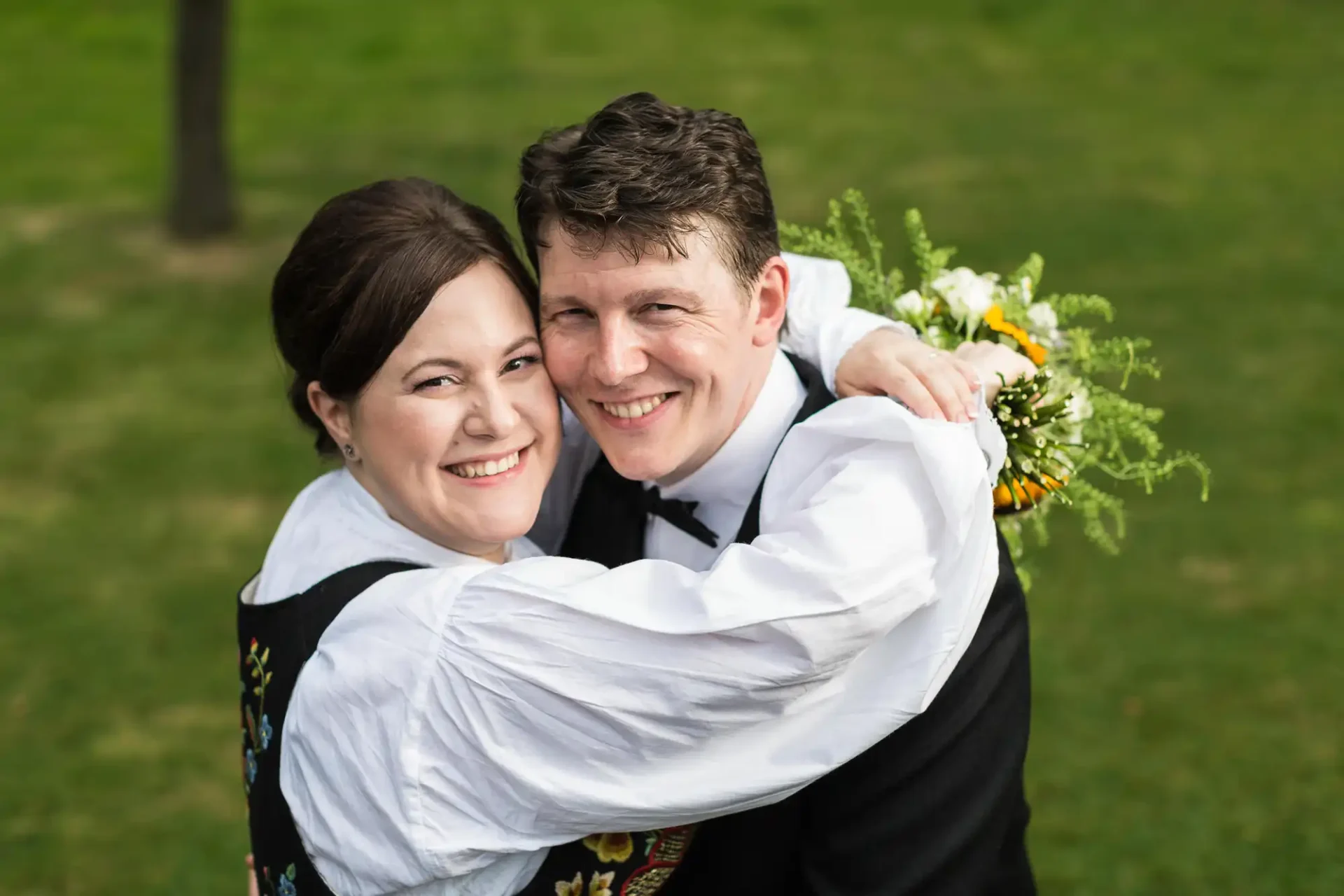 A joyful couple in traditional attire embracing, with the woman holding a bouquet, outdoors on a grassy background.