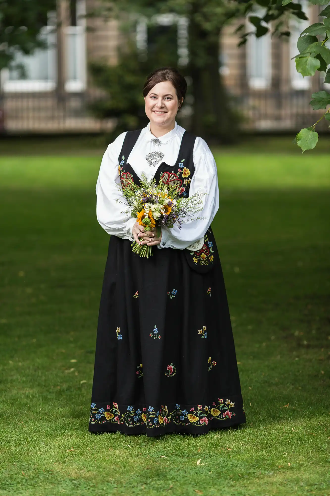 Woman in traditional norwegian bunad with embroidered details, holding a bouquet, smiling in a garden.