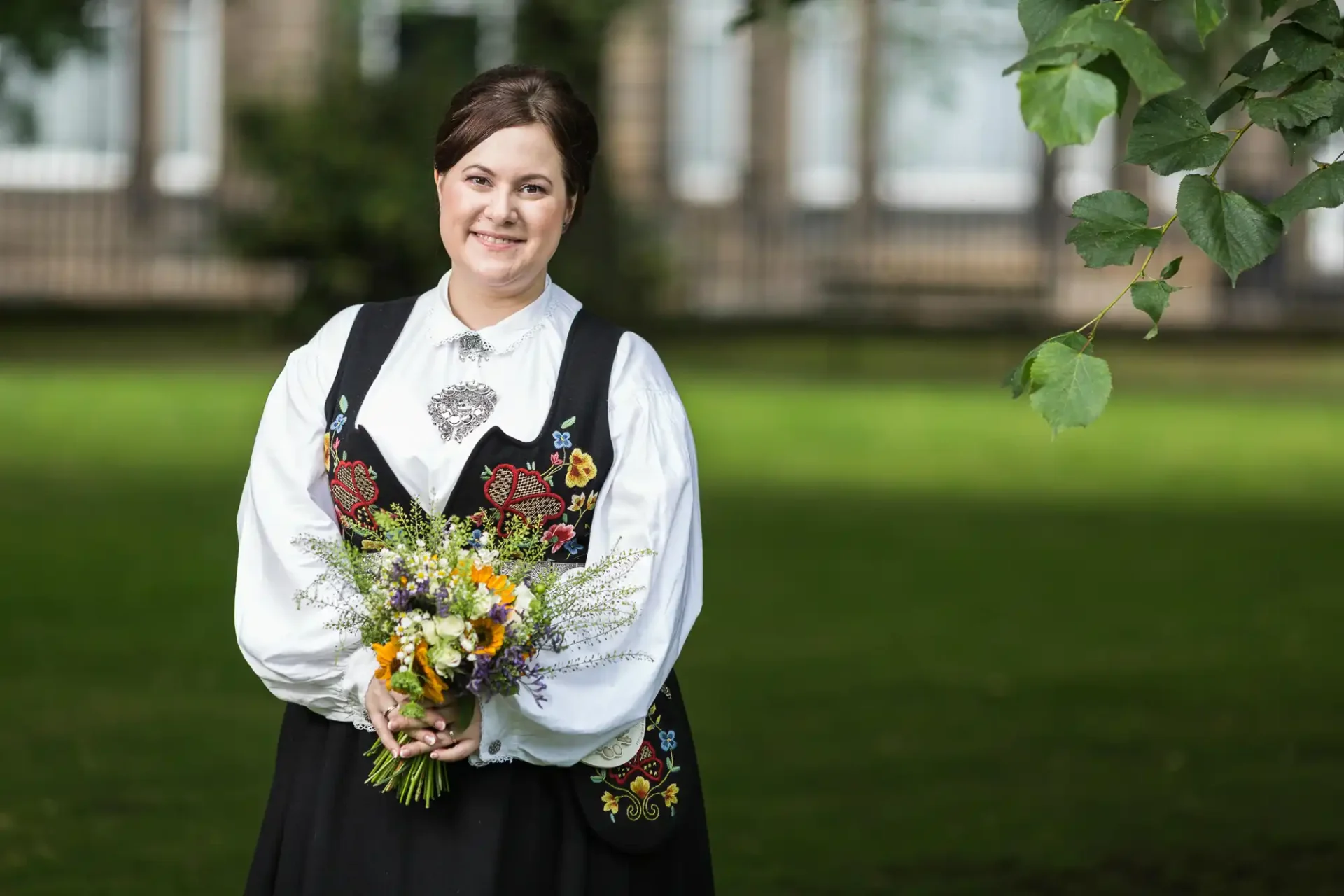 A woman in a traditional norwegian bunad, smiling and holding a bouquet of wildflowers, standing in a park.