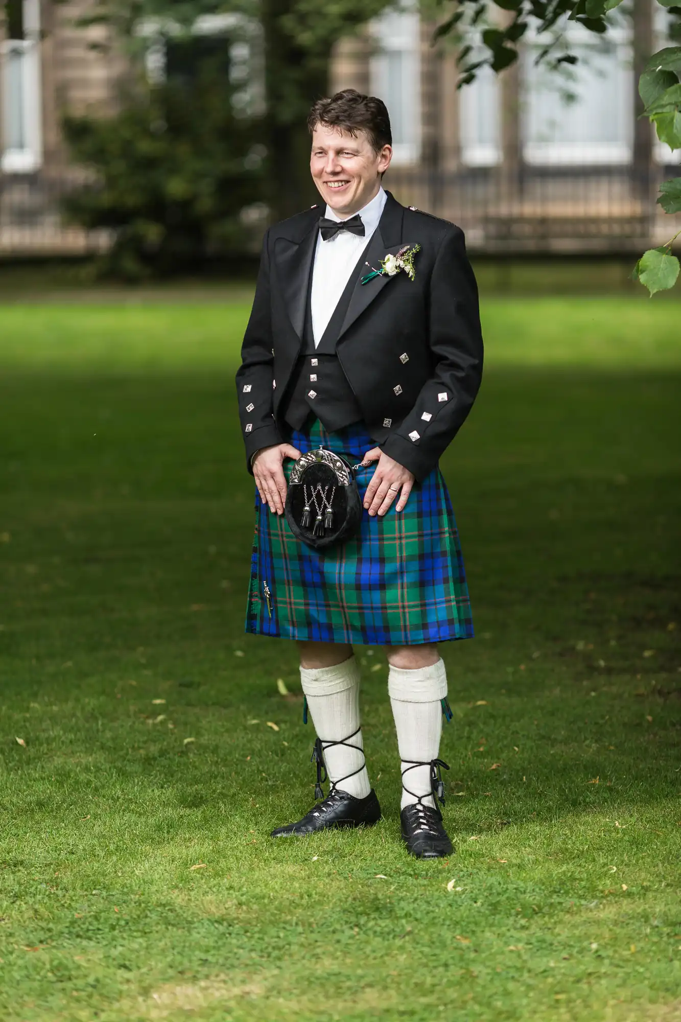 A person standing on a grassy lawn wearing a traditional scottish outfit, including a kilt with a tartan pattern, sporran, and black jacket.