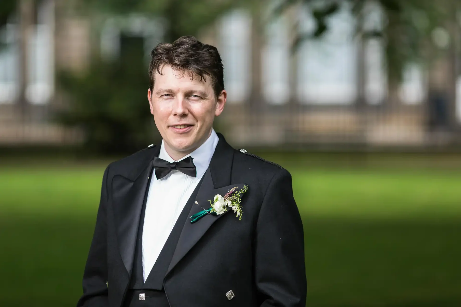 A man in a tuxedo with a bow tie and boutonniere smiling in a park setting.