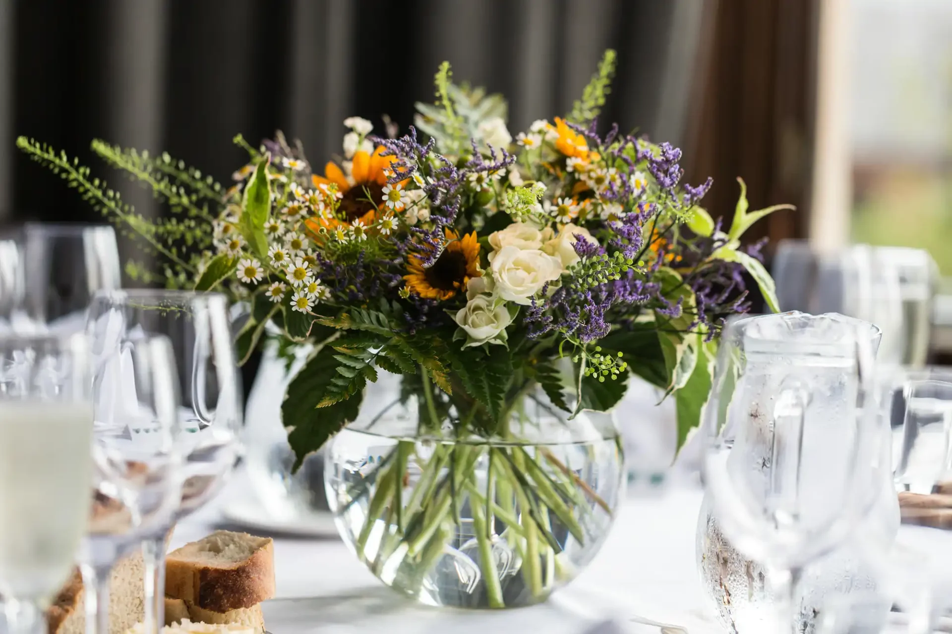 Elegant floral centerpiece in a glass vase with orange, yellow, and purple flowers on a dining table with glasses and bread.