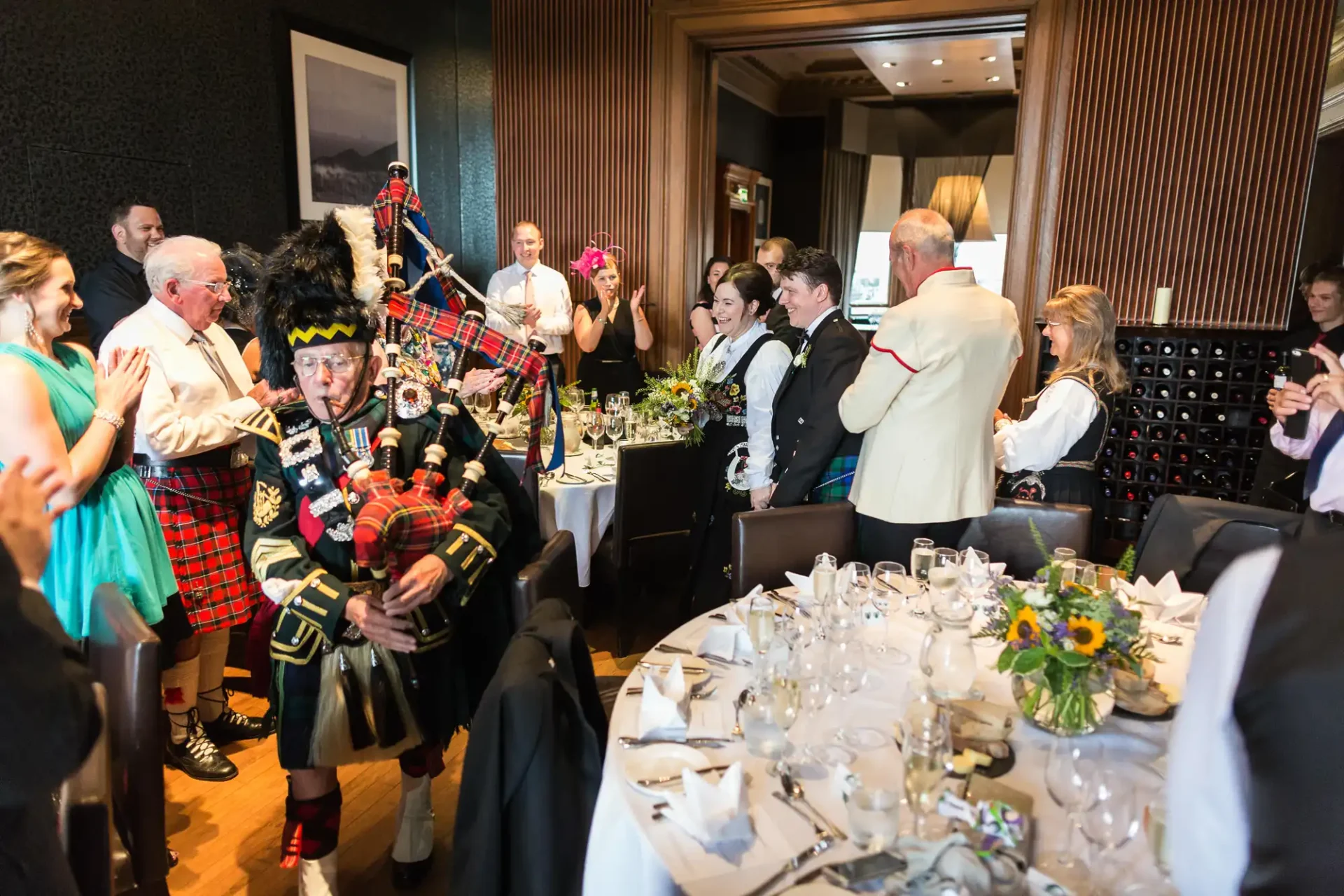 A man in traditional scottish attire playing bagpipes at a lively event inside a dining room with guests enjoying the performance.