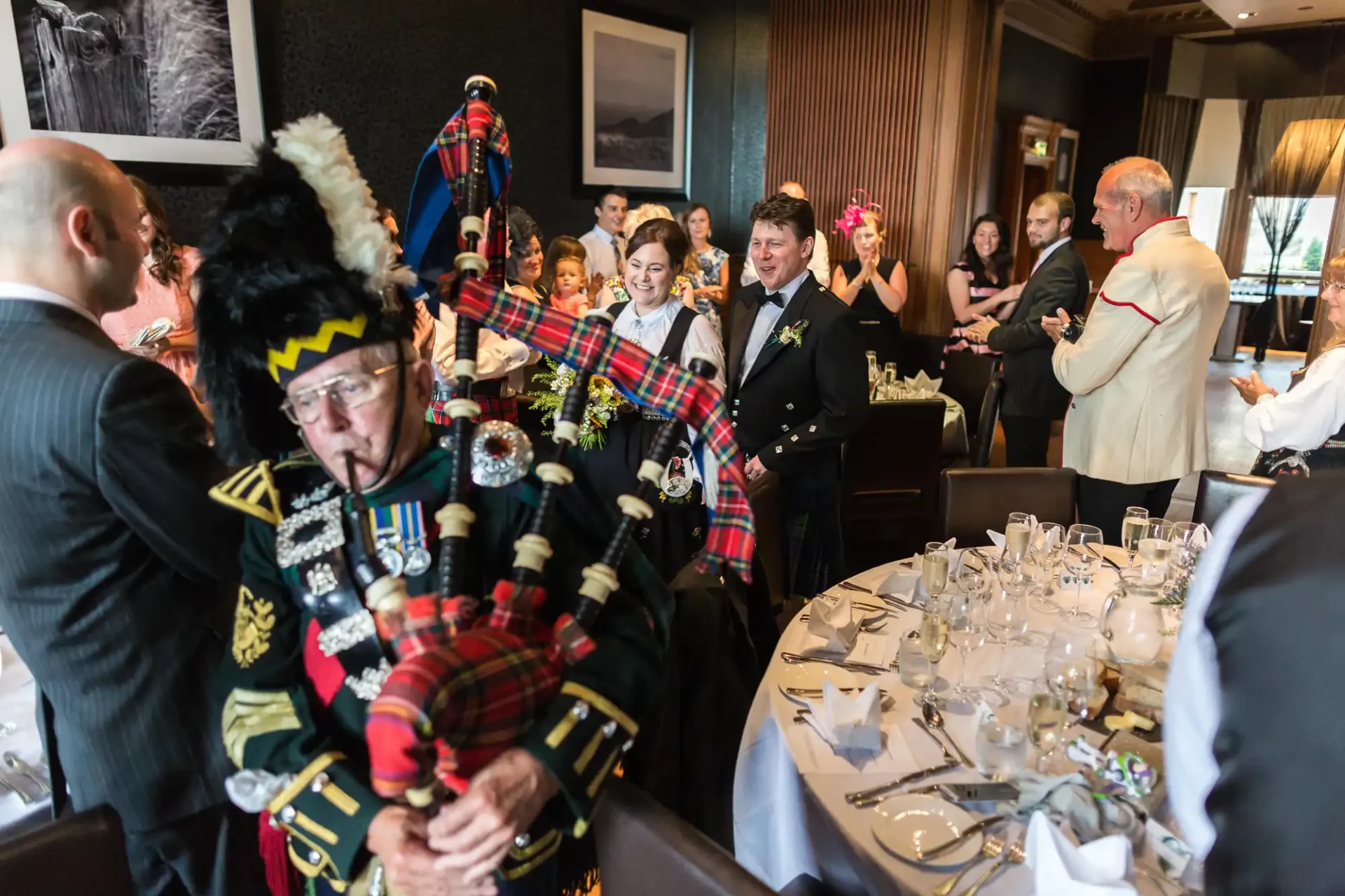 A bride and groom follow a bagpiper in traditional scottish attire through a festively decorated dining room at their wedding reception.