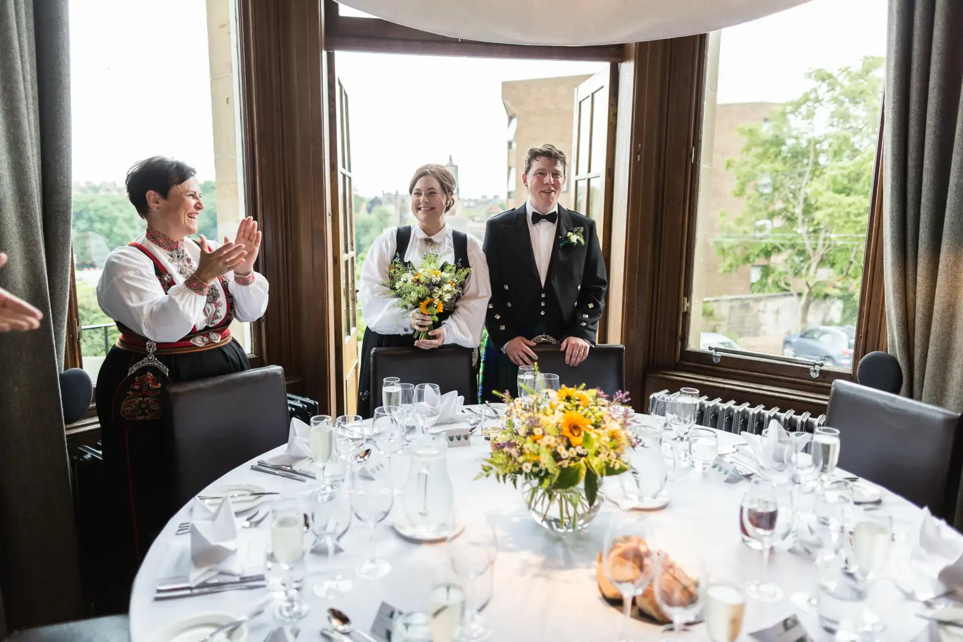 A wedding couple stands smiling at a decorated table in a restaurant, with a guest clapping and a city view through large windows.