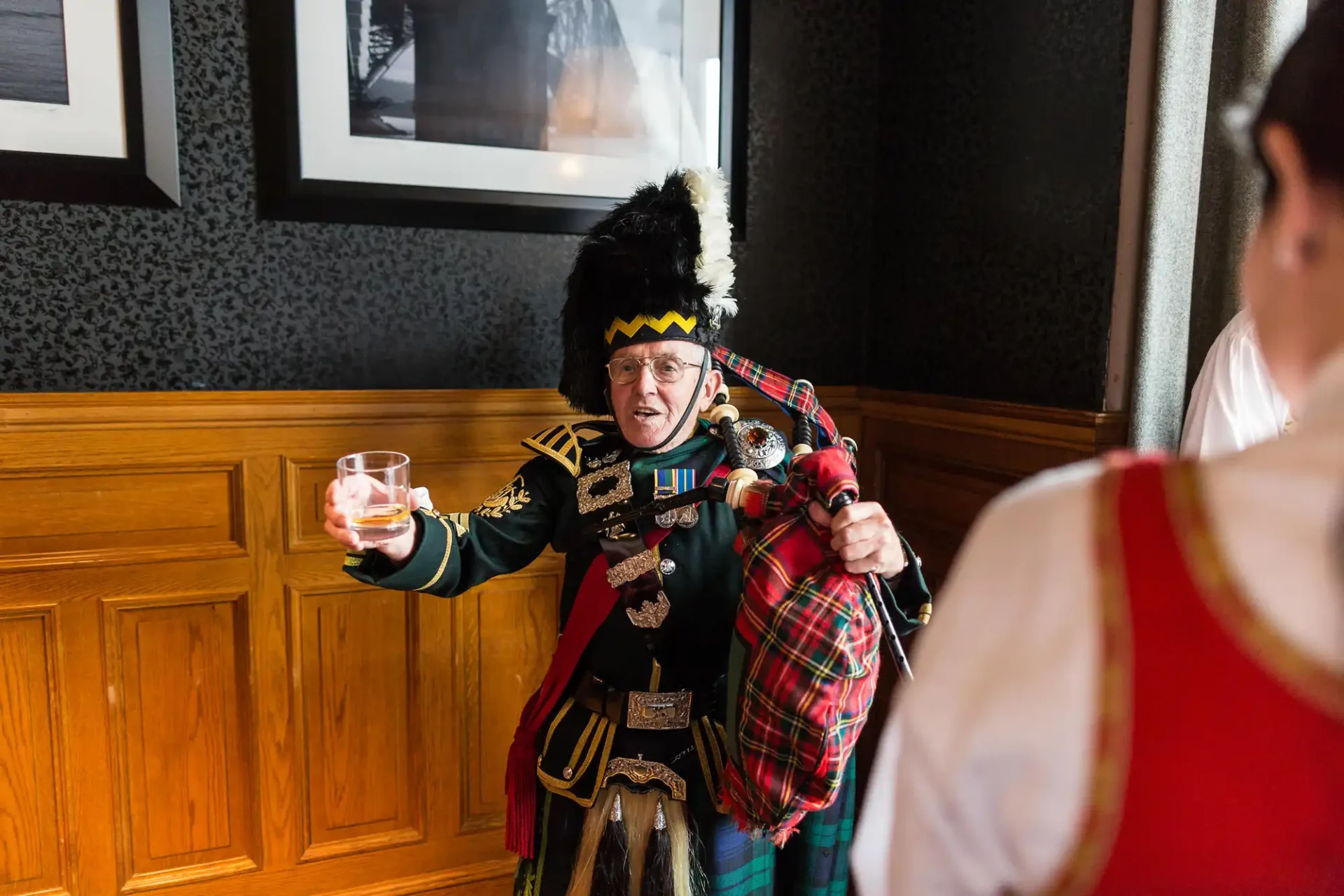Man in traditional scottish attire, including kilt and sporran, holding a glass of whiskey, in a room with wood-paneled walls and framed photos.