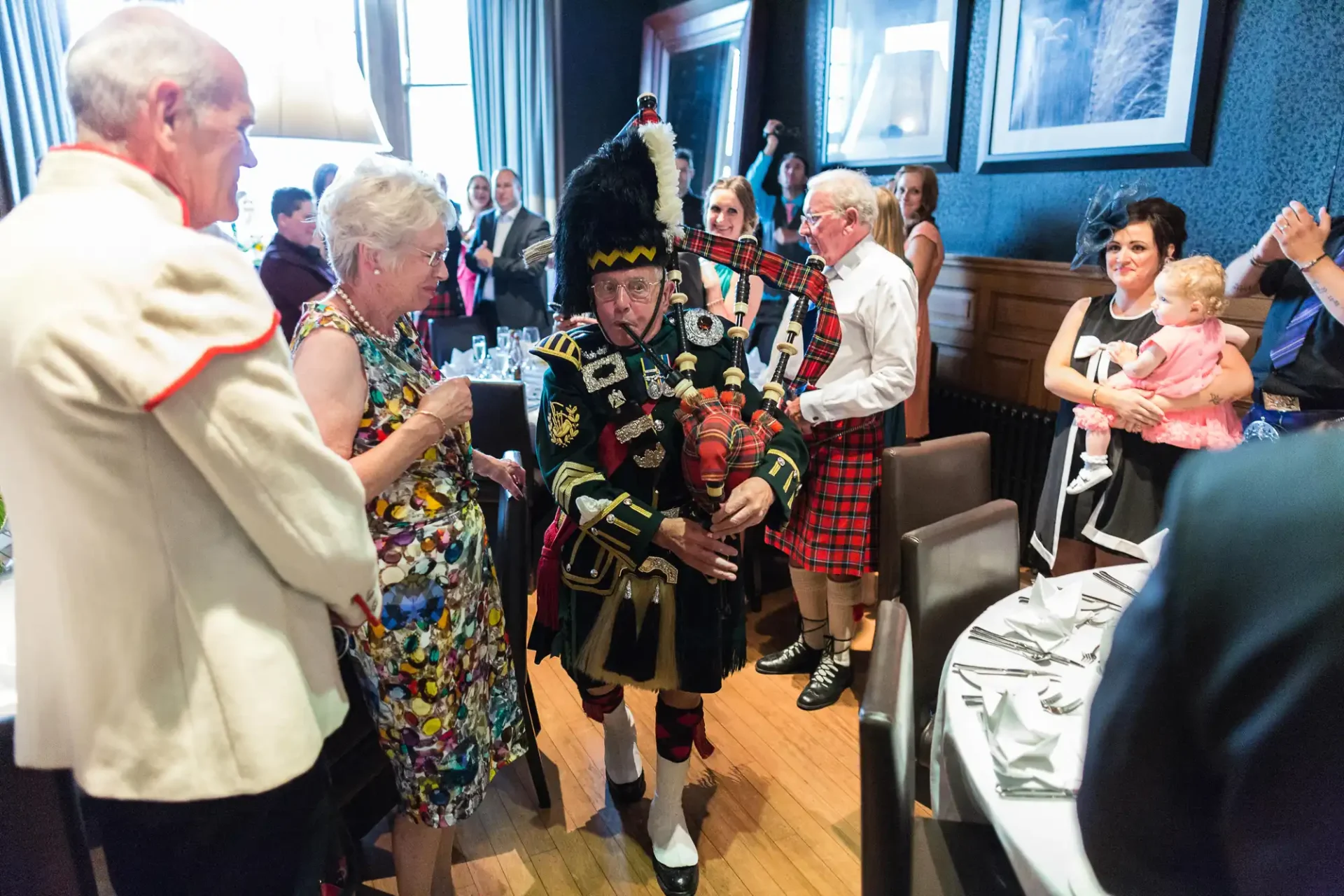A bagpiper in traditional scottish attire playing at a lively indoor event with guests in casual and formal wear watching and interacting.