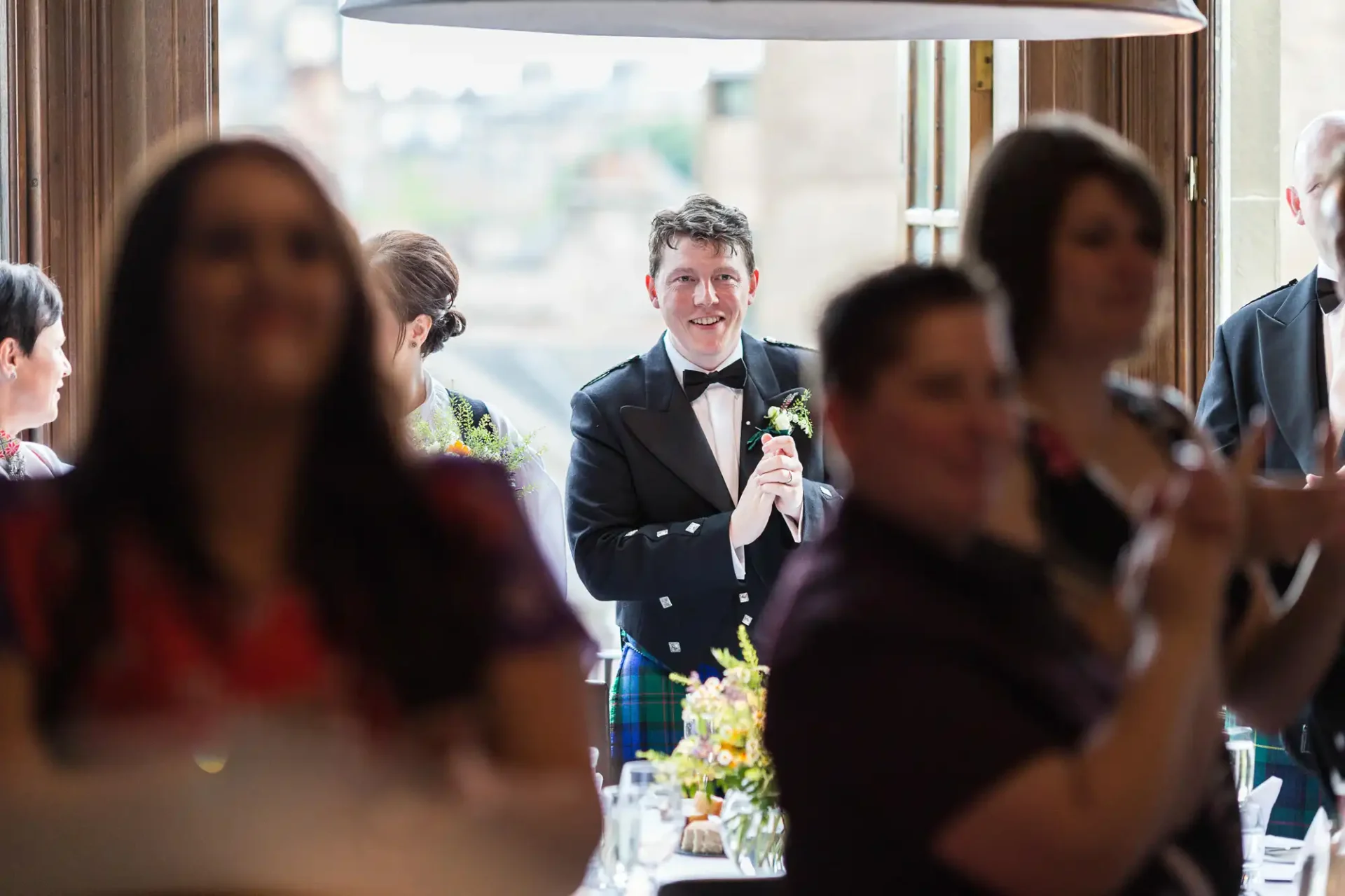 Man in a kilt smiling at a wedding reception, standing between blurred foreground figures and a bright window.