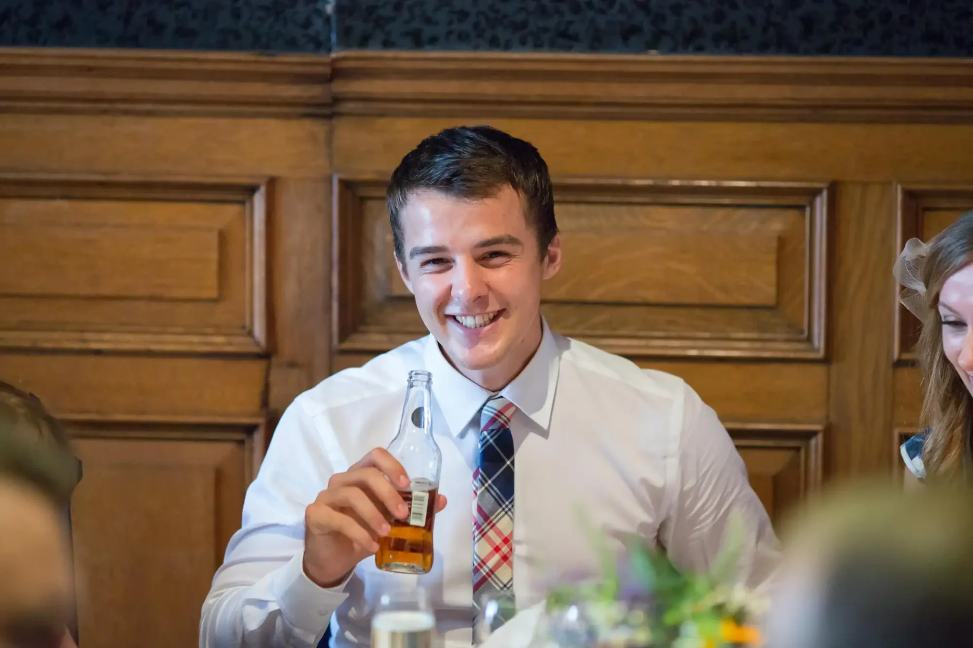 A smiling young man in a white shirt and plaid tie holding a bottle of beer at a formal event.