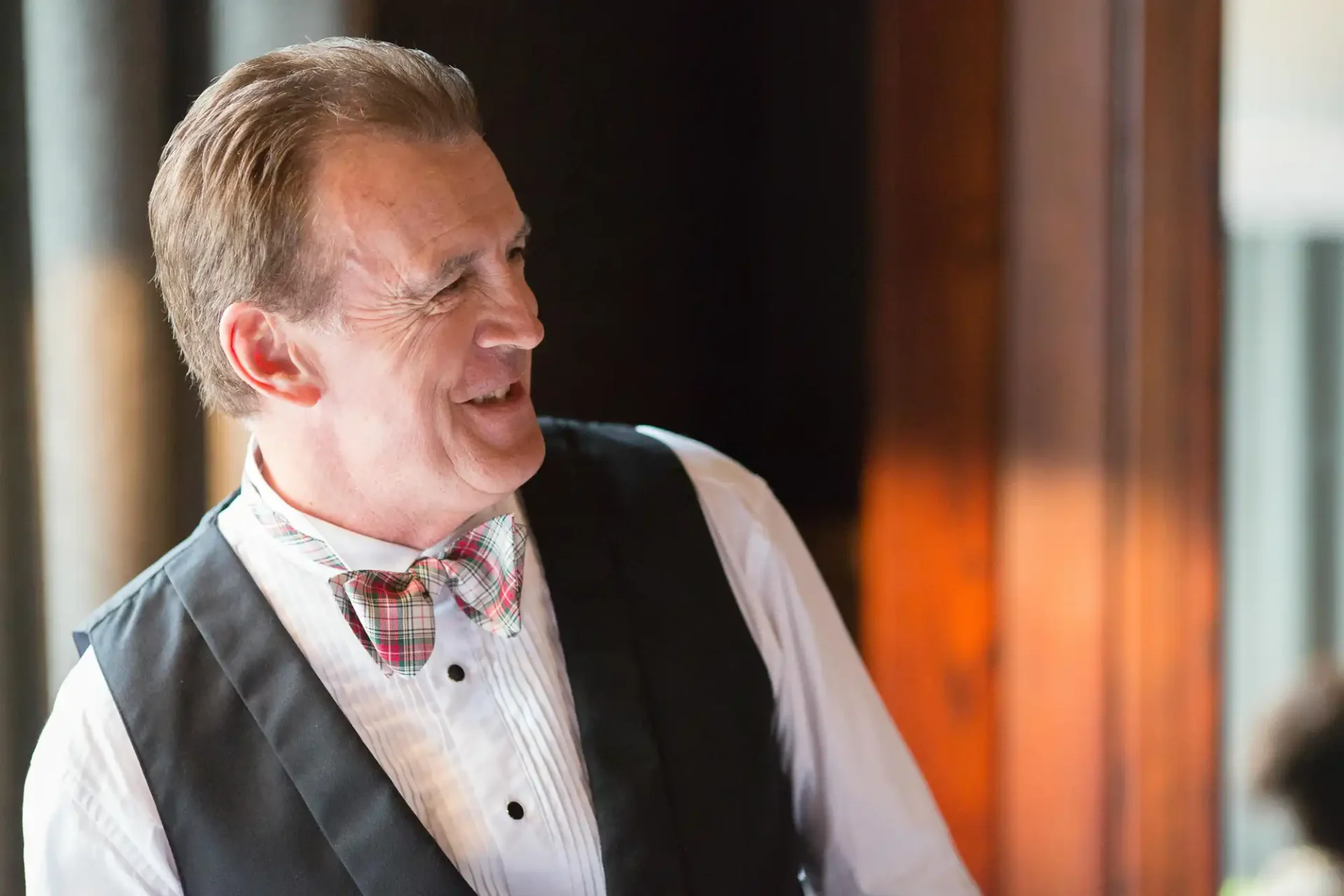 Man in a vest and bow tie smiling joyfully in a warmly lit room.