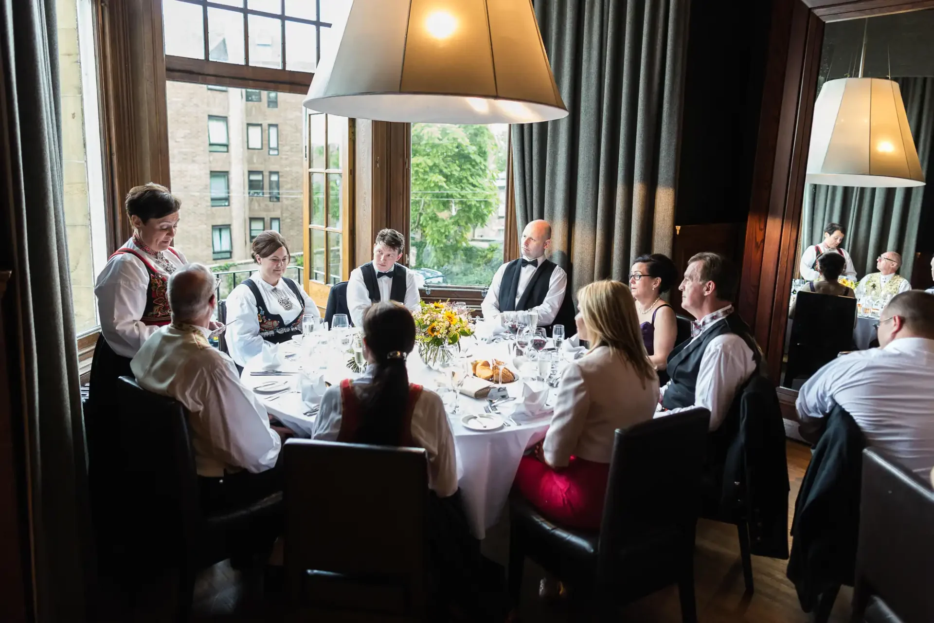 Waitstaff serving a formally dressed group at an elegant dining table in a restaurant with large windows and modern decor.