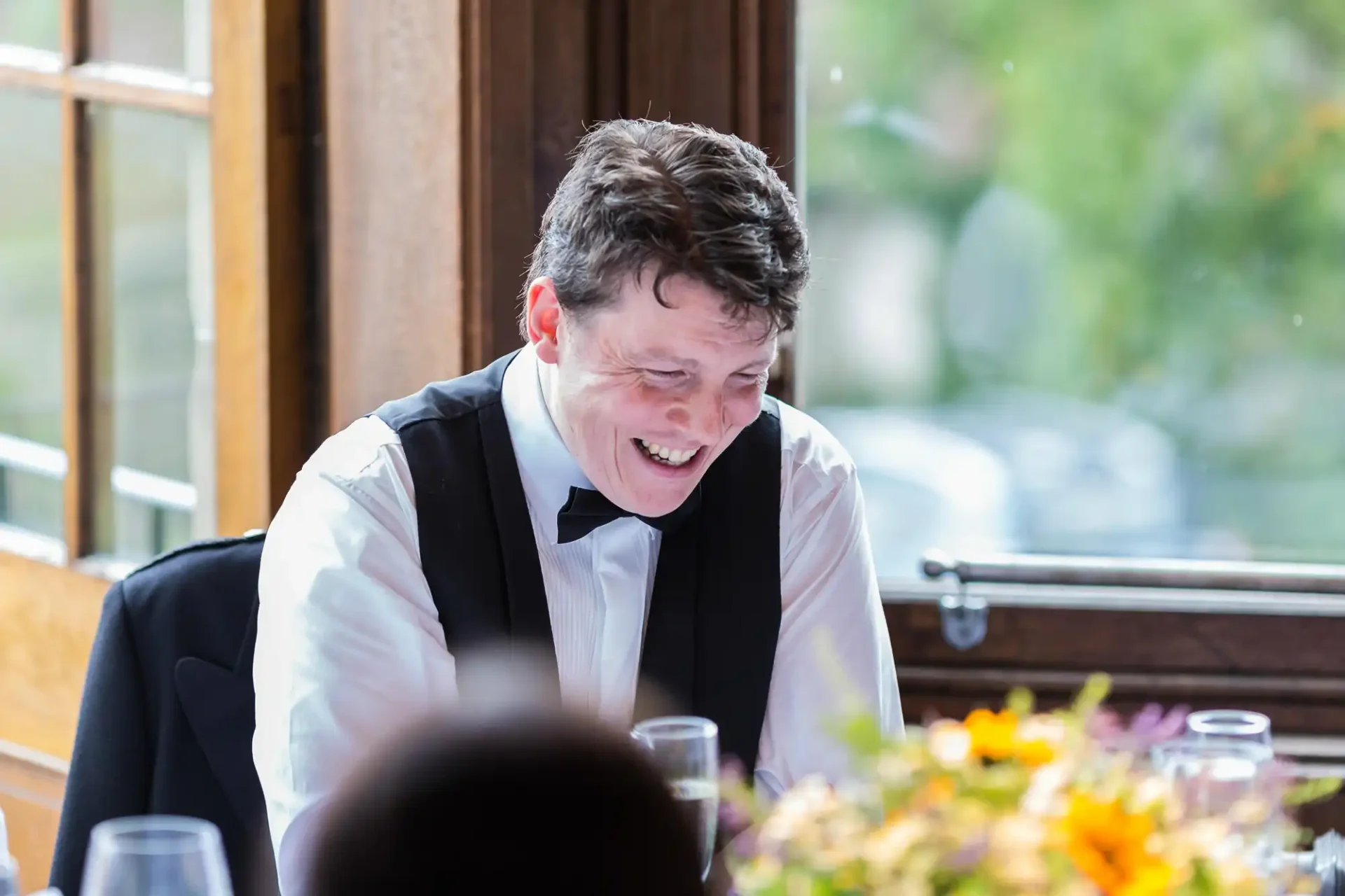 A man in a tuxedo laughing joyfully at a table with a floral centerpiece, natural light streaming through a window beside him.