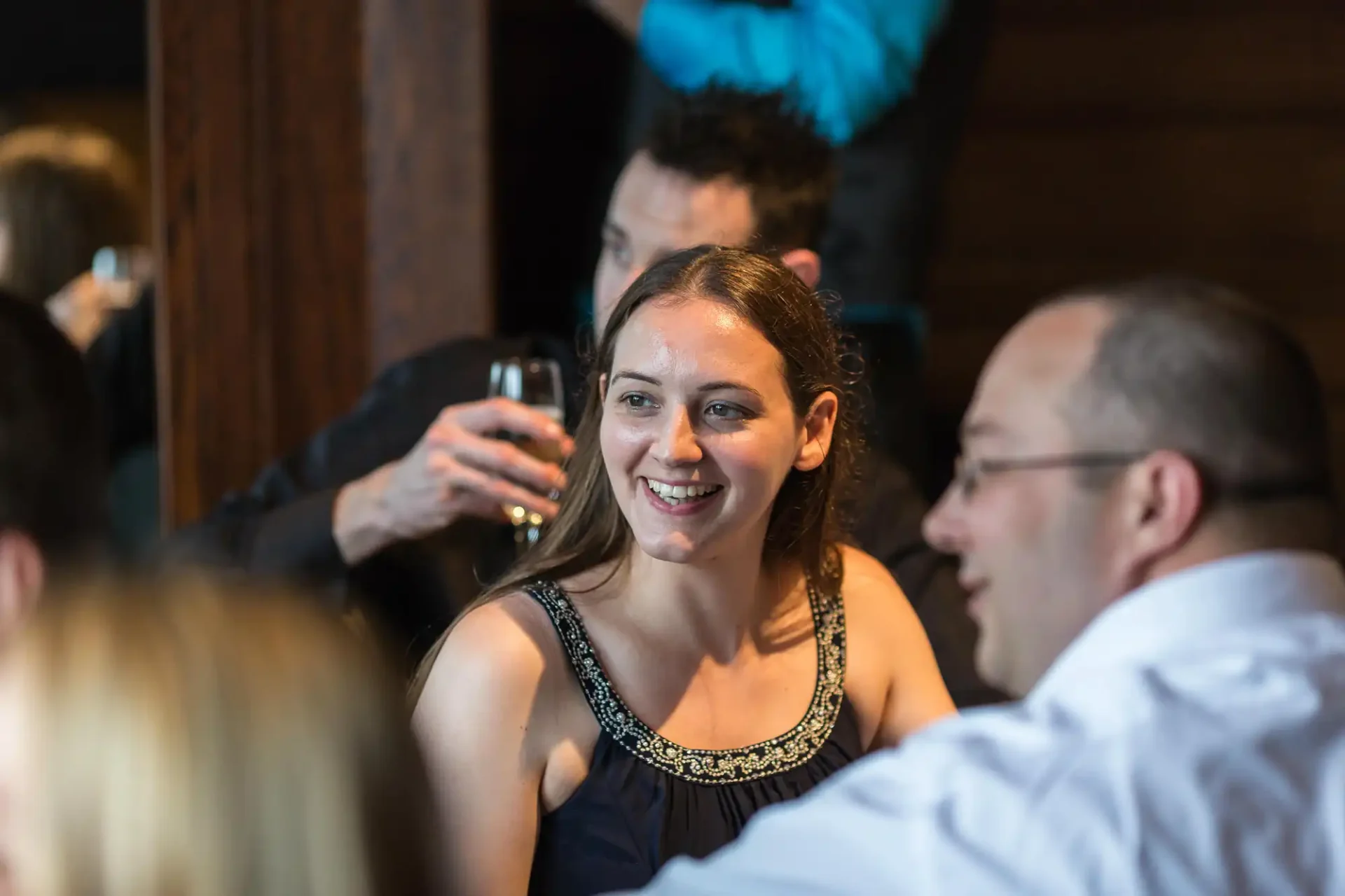 A woman in a black dress smiles during a conversation with two men at a social event, with another person holding a drink in the background.
